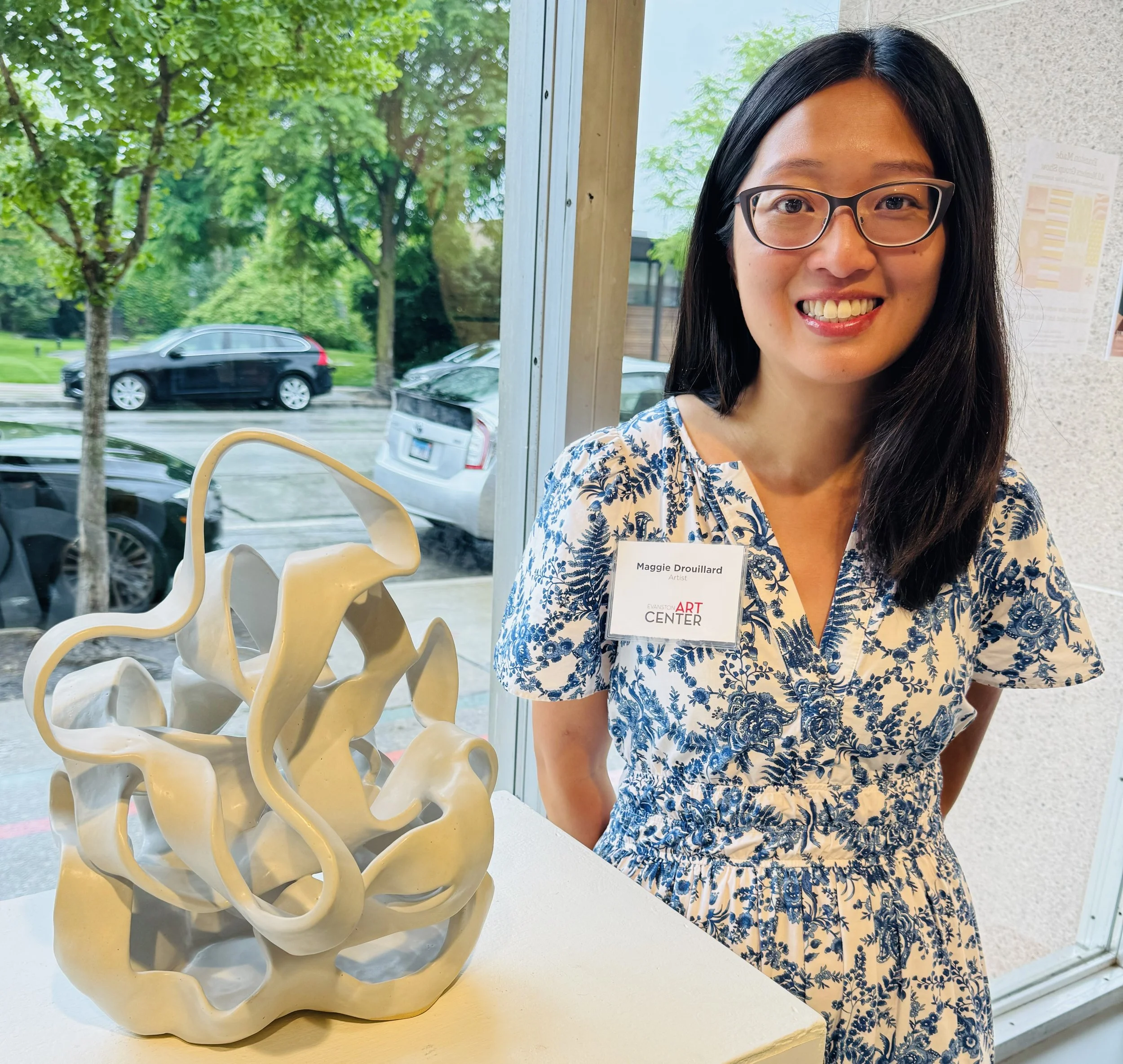 A woman with glasses wearing a blue and white floral dress standing next to a ceramic art sculpture inside an art center, with a window showing parked cars and green trees outside.