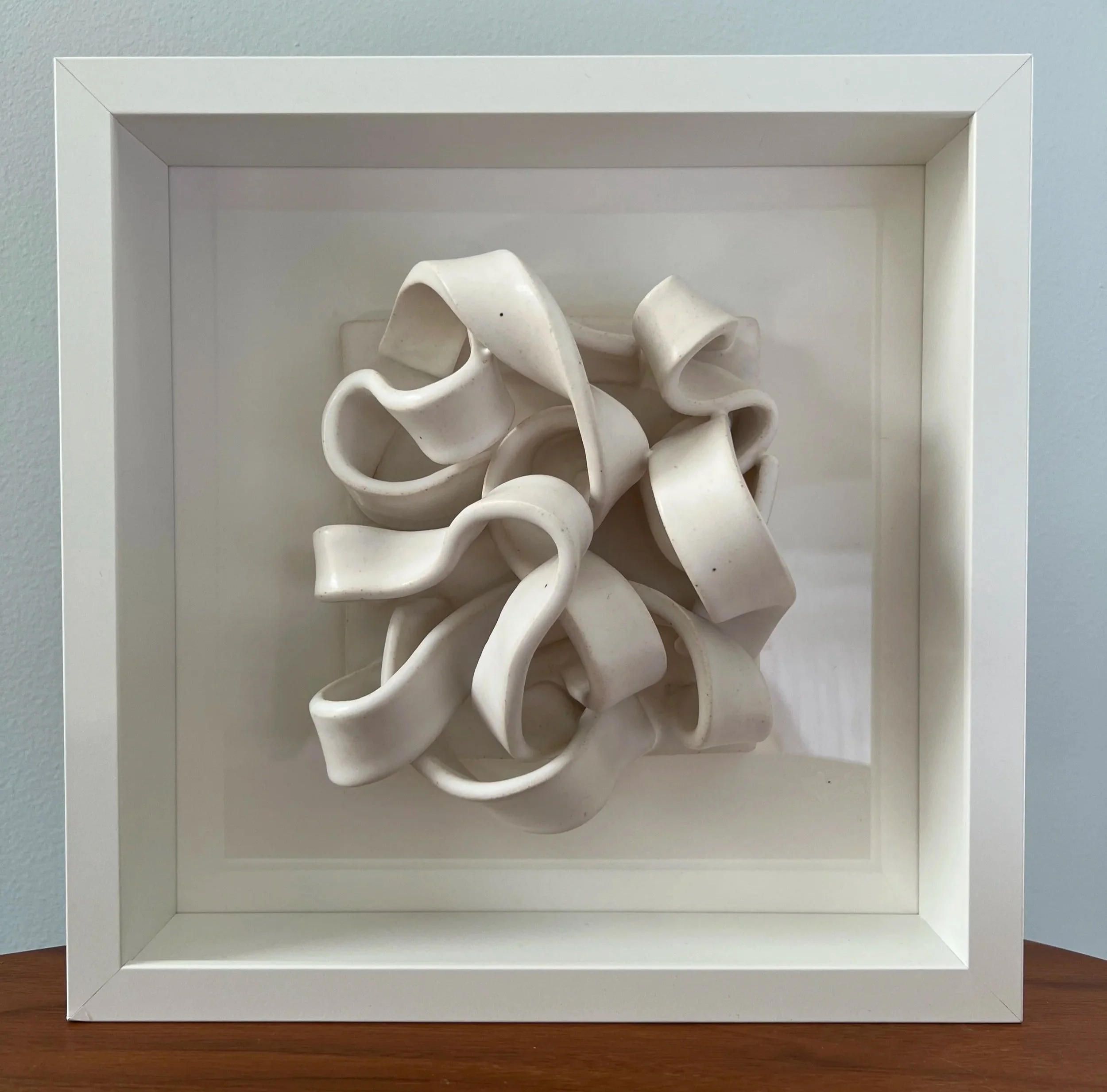 White ceramic sculpture arranged in a shadow box frame, with the sculpture consisting of ribbon-like loops and twists.