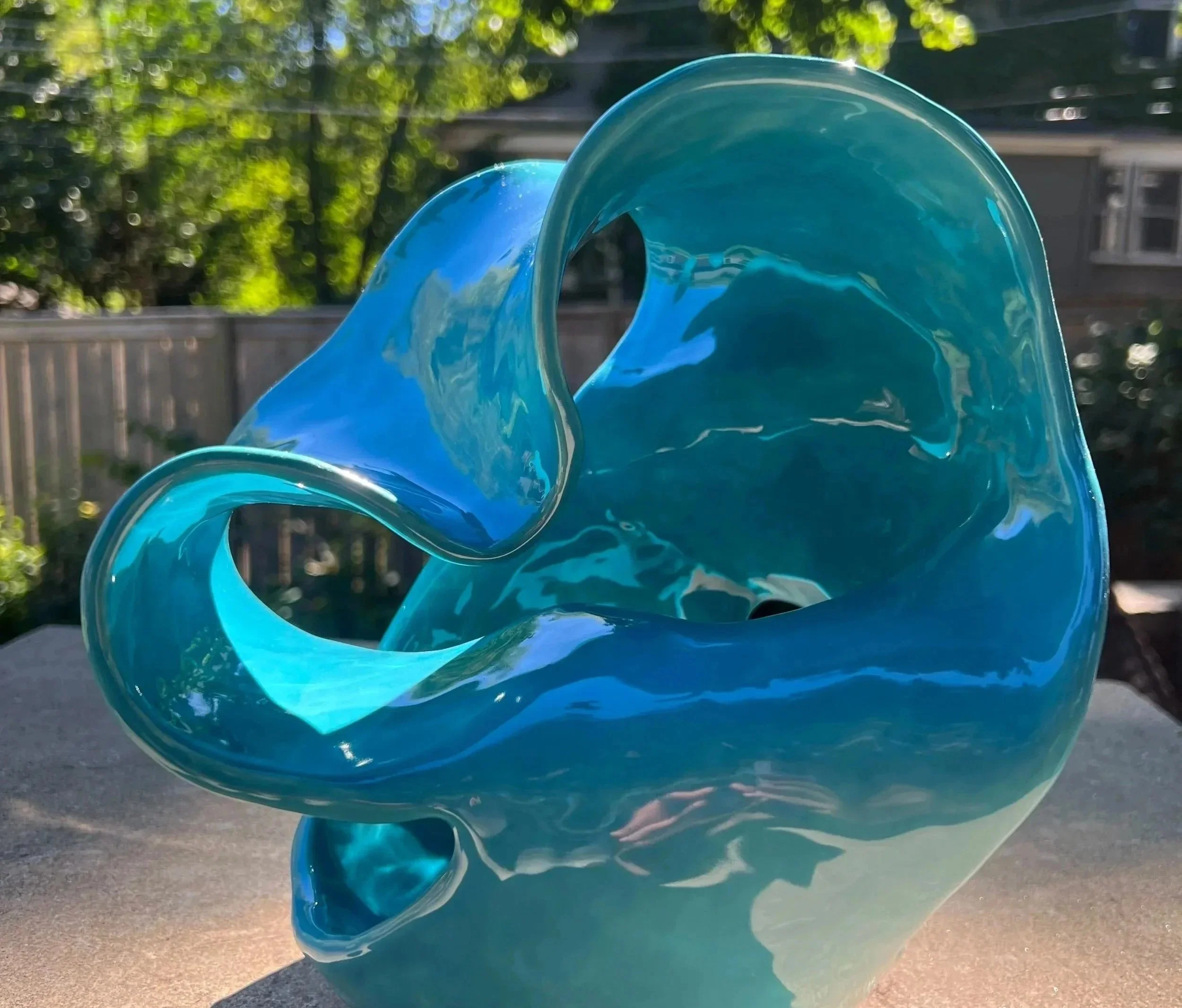 A glass sculpture with flowing, curving shapes in shades of blue, set outdoors with trees and a building in the background.