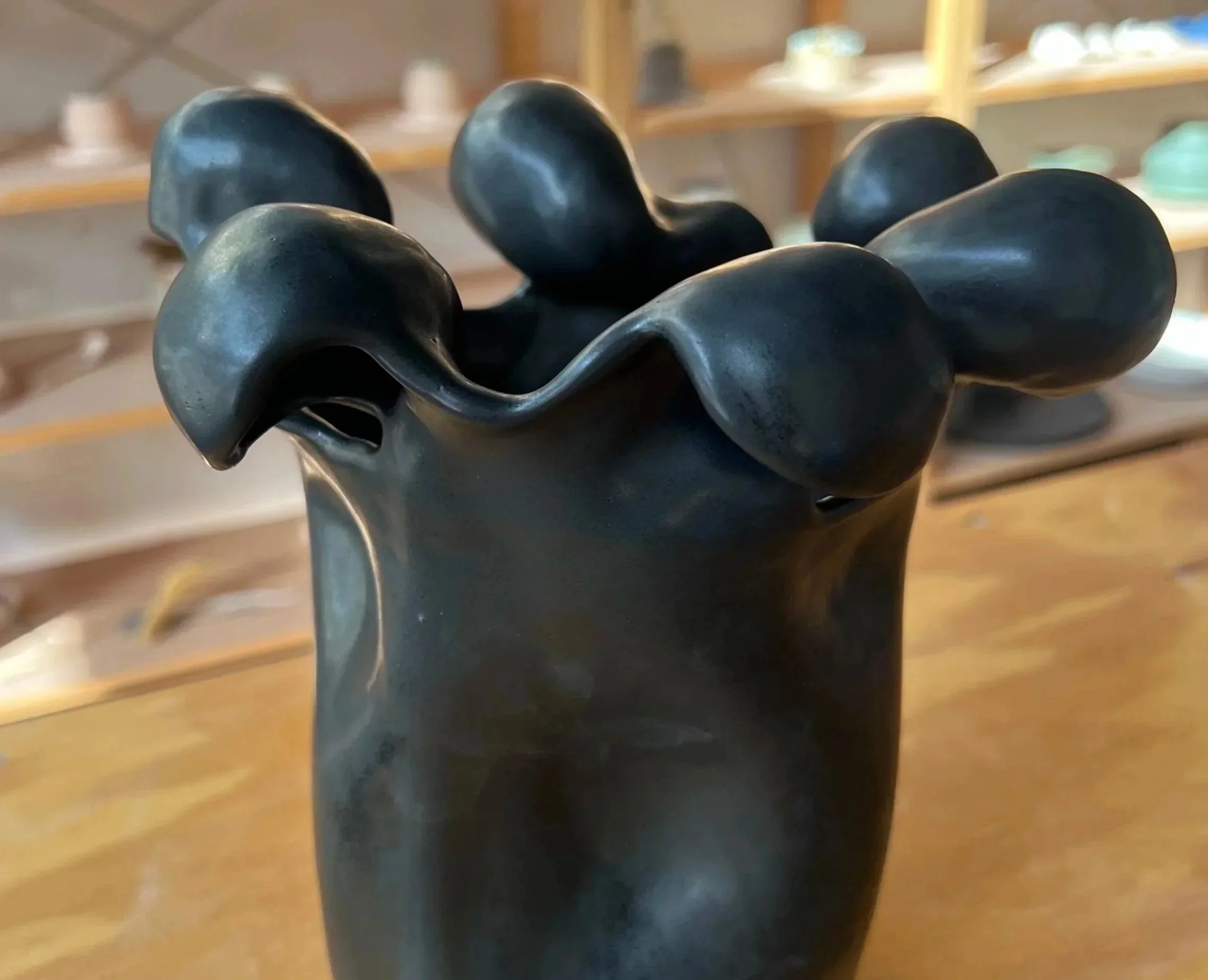 Close-up of a black ceramic sculpture resembling a vase with abstract, flowing shapes on the top, placed on a wooden surface with shelves in the background.