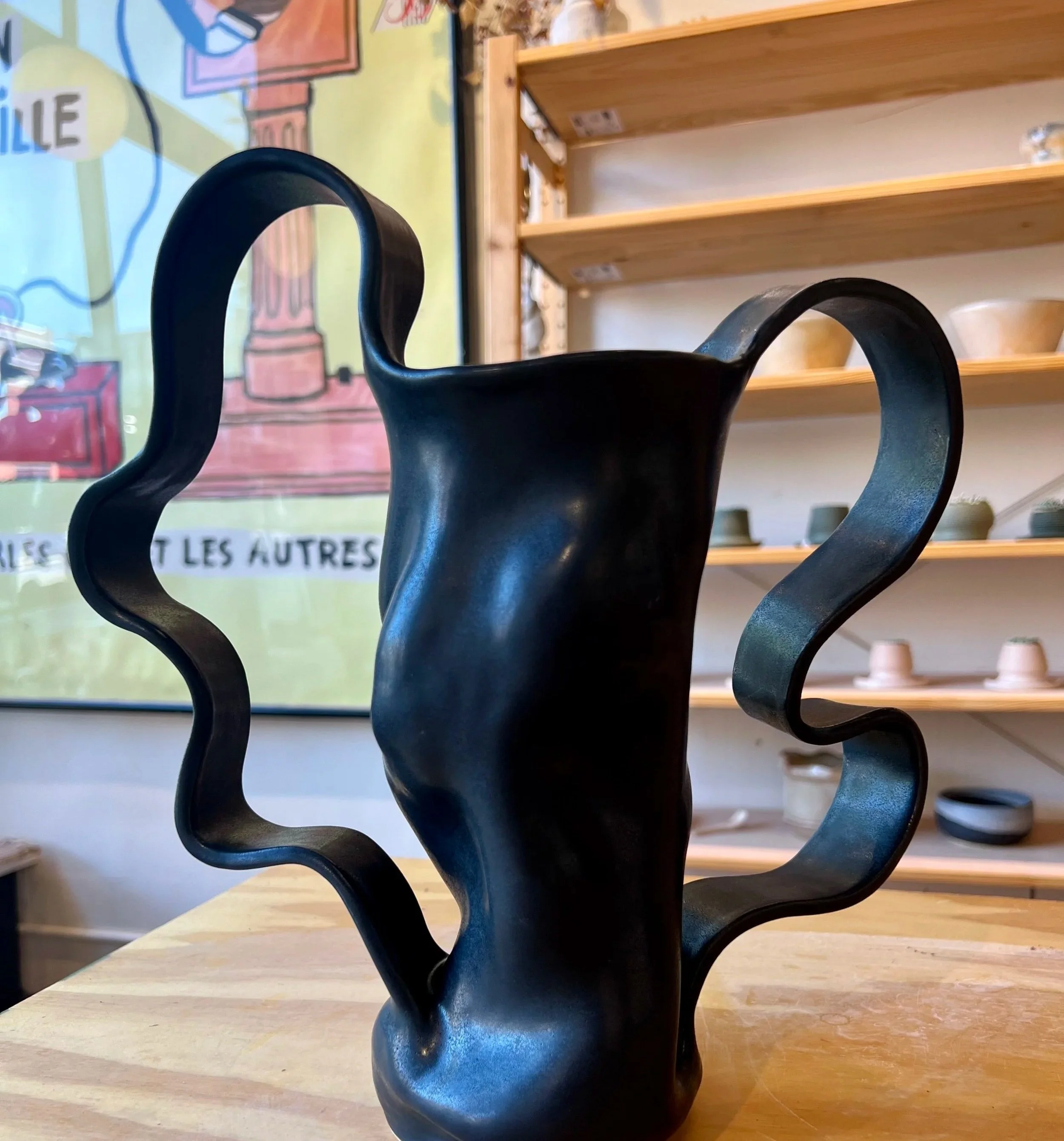 A black ceramic vase with wavy handles on display in a store, with wooden shelves holding various ceramic pieces in the background.
