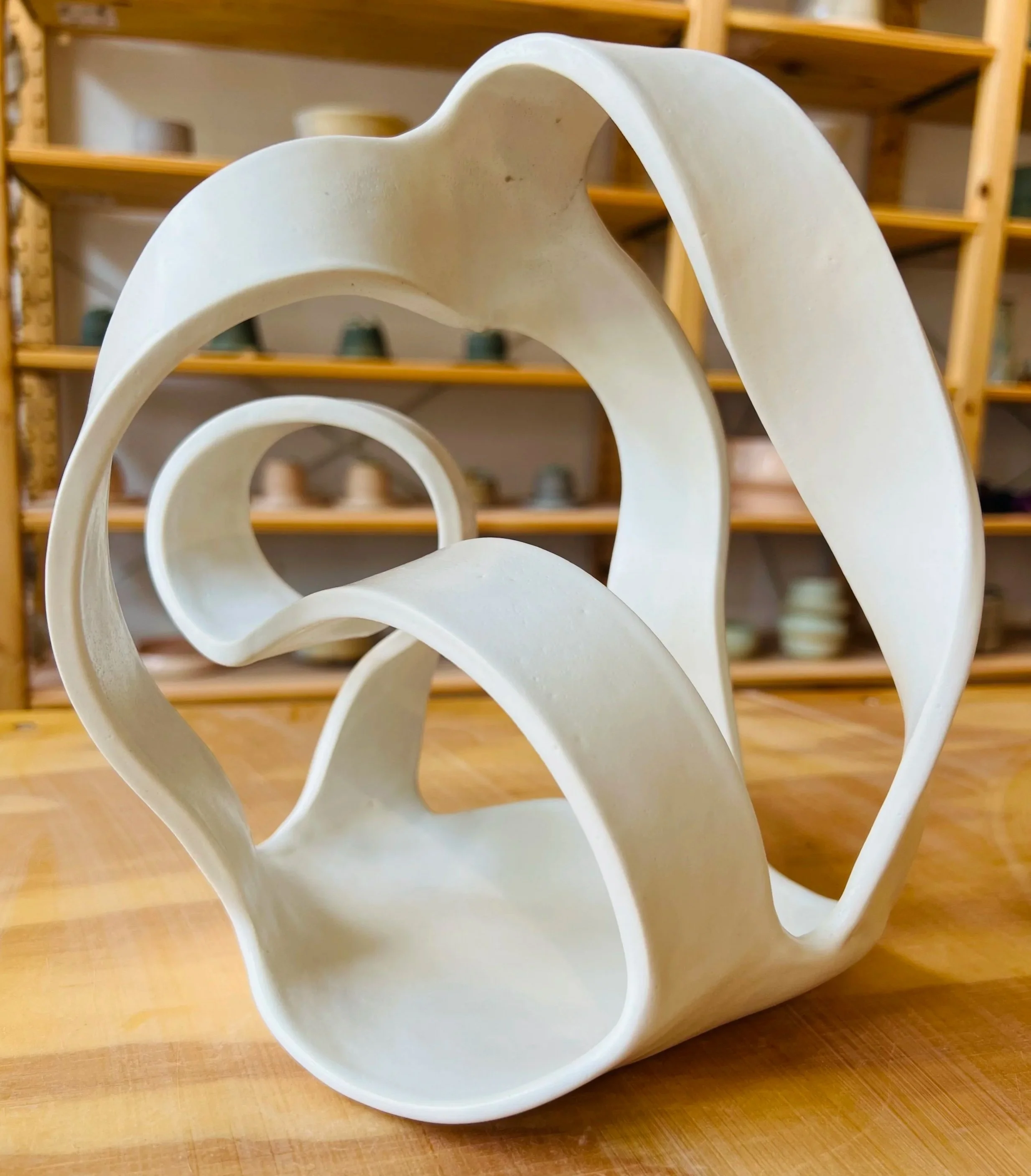 A white, abstract ceramic sculpture with flowing, curved lines, placed on a wooden surface with shelves of various ceramic bowls and cups in the background.