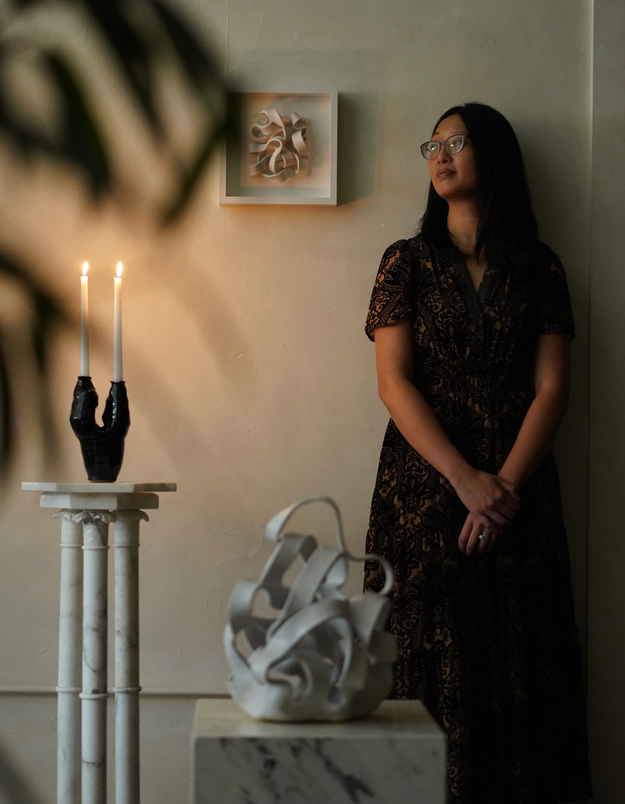 A woman with black hair and glasses standing against a beige wall, wearing a black lace dress, with her hands clasped in front of her. There is a small white table with a decorative sculpture in the foreground, and a candle holder with two lit candles on another stand nearby. A framed abstract art piece is hanging on the wall.