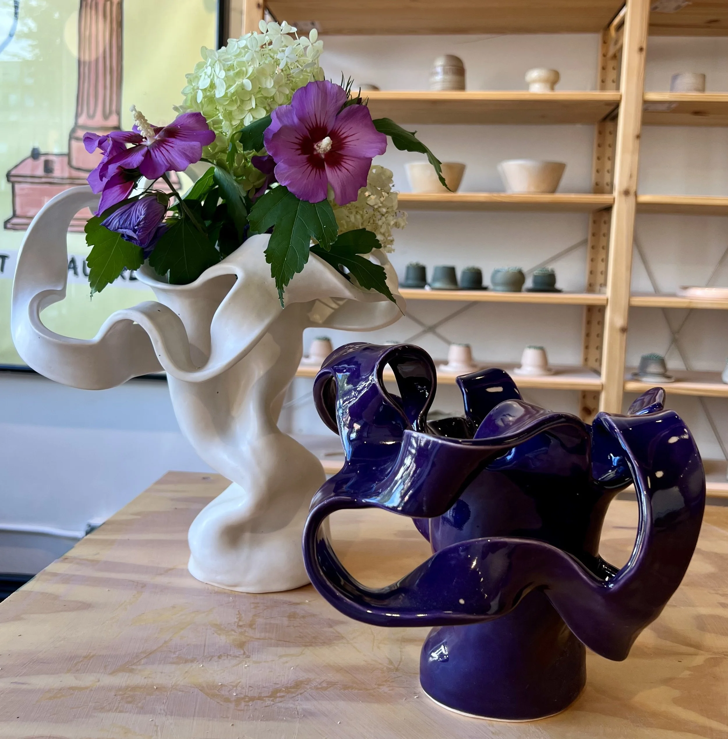 A white, abstract ceramic vase with a wavy, flowing design holding a bouquet of purple and white flowers, next to a dark purple ceramic sculpture with a similar wavy, flowing design, set on a wooden table in a pottery studio with shelves of bowls and cups in the background.