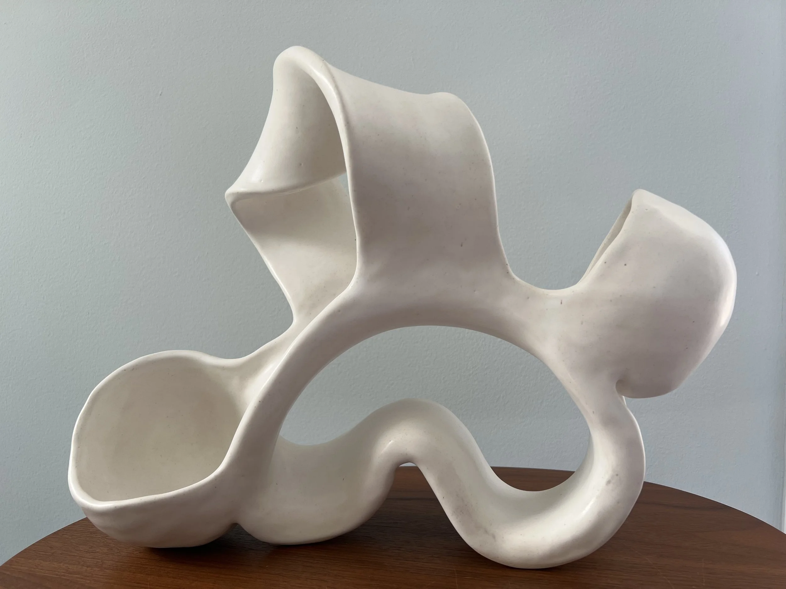 White abstract ceramic sculpture with flowing curves on a wooden surface against a light blue wall.