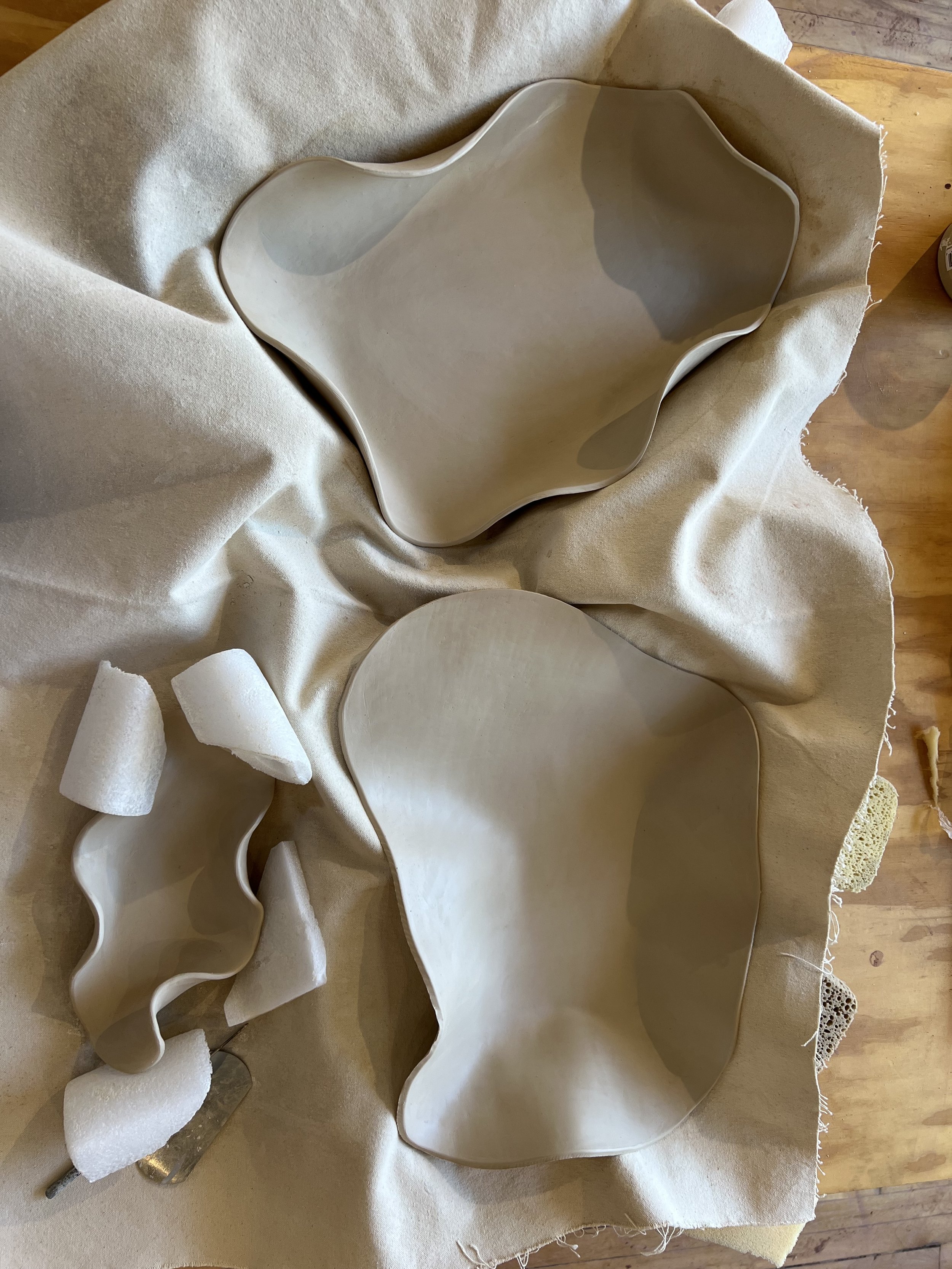 Two porcelain bowls with wavy edges resting on fabric, with foam padding and tools nearby on a wooden surface.