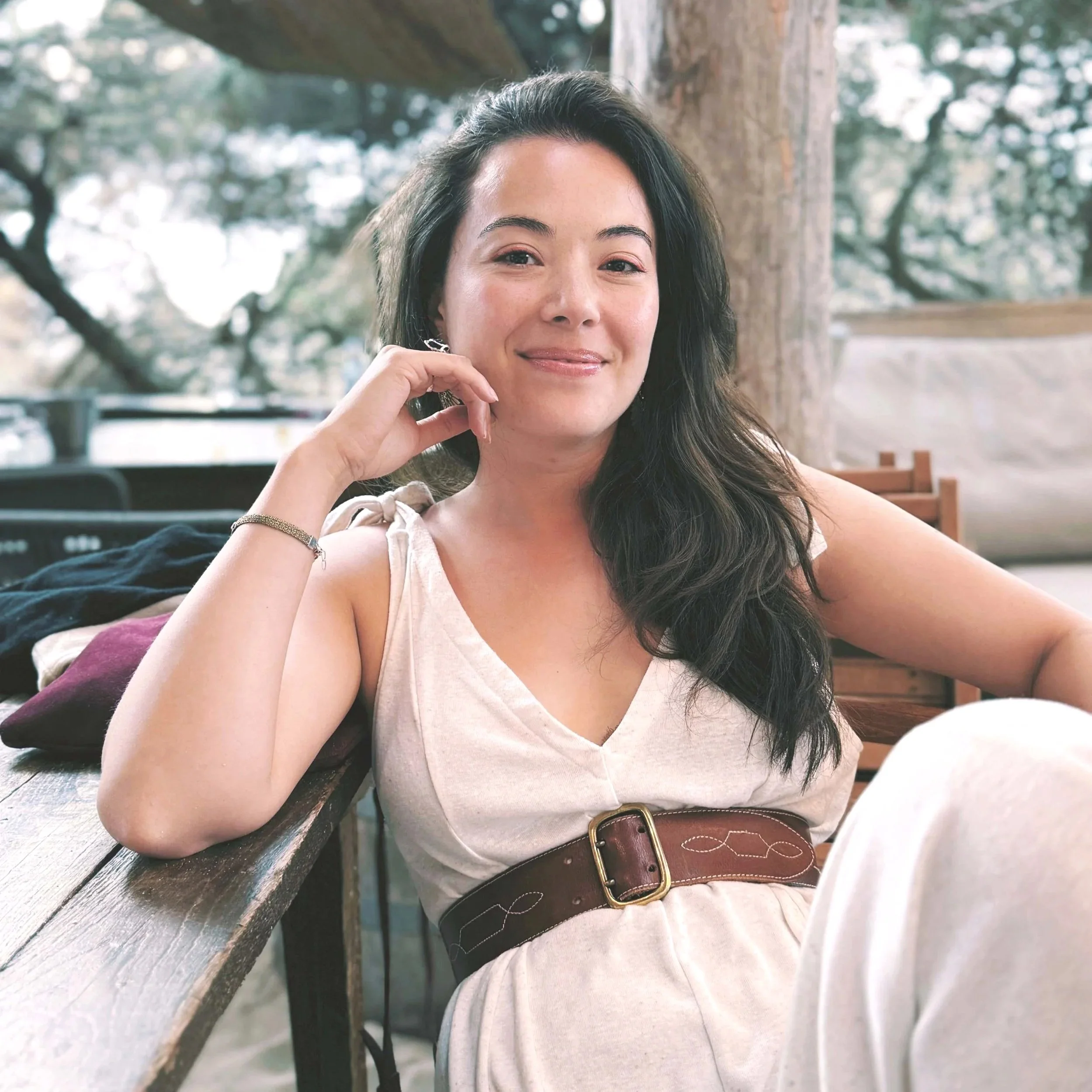 A woman with long dark hair, wearing a white dress and jewelry, sitting outdoors at a wooden table with a blurred green and tree background.