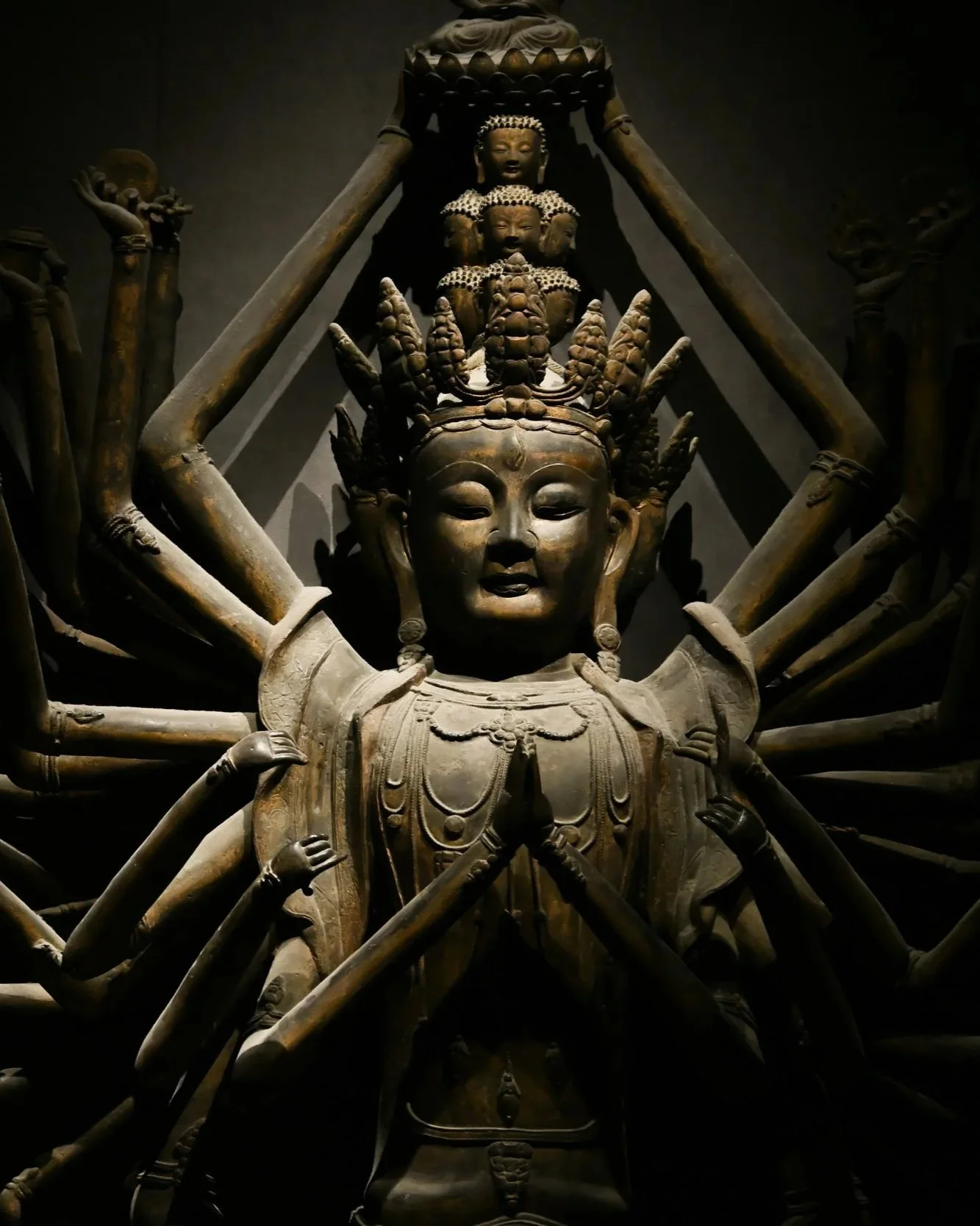 A large bronze sculpture of a deity with multiple arms and a serene facial expression, adorned with elaborate headdresses and jewelry, in a dimly lit setting.