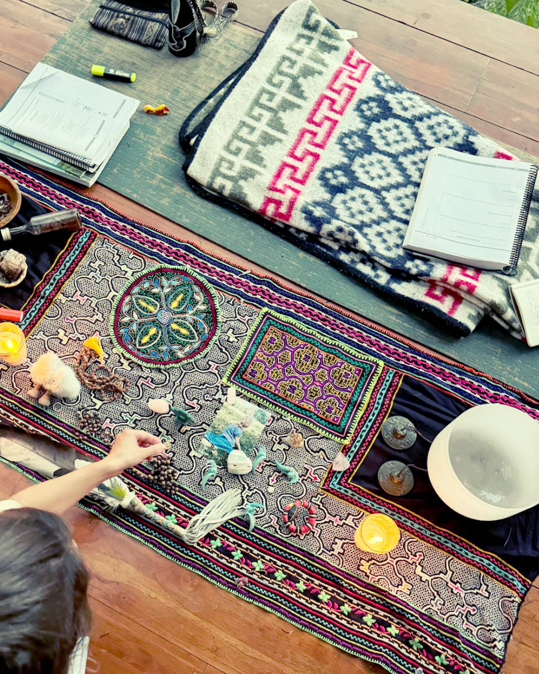Colorful embroidered textile with beads and small decorations, placed on a wooden floor, with candles and other objects around, and a person reaching toward it.