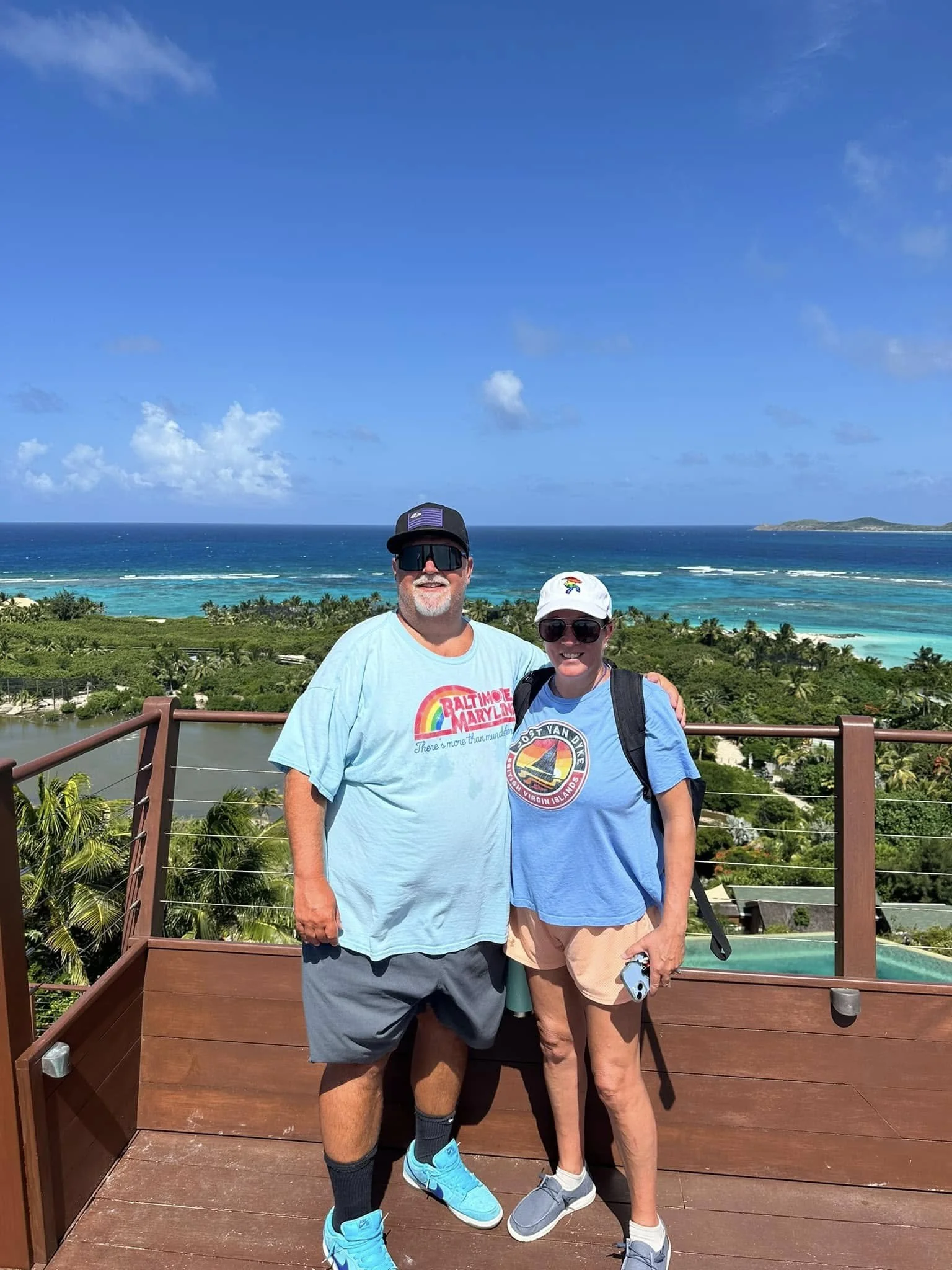 Two people standing on a wooden observation deck with a scenic ocean view behind them. The man is wearing a light blue t-shirt, shorts, and blue sneakers, and the woman is wearing a blue t-shirt, shorts, and gray sneakers. Both are wearing sunglasses and hats.