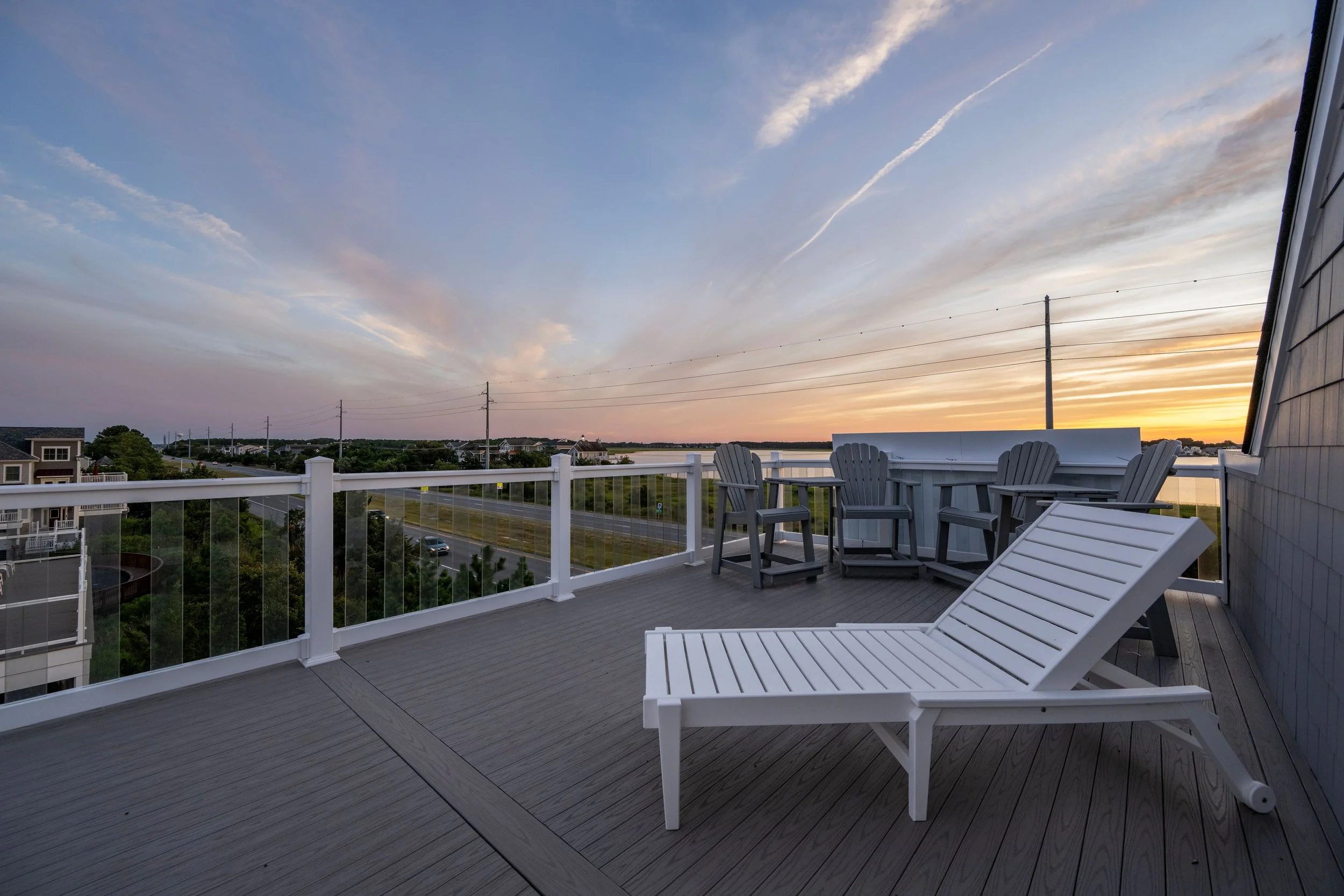 Empty outdoor balcony with white lounge chair and Adirondack chairs at sunset, overlooking a waterway and distant houses.