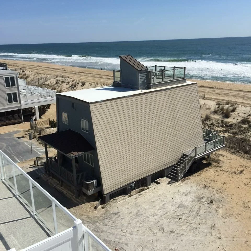 A house on stilts with a deck on top, facing the ocean, with stairs leading down to a sandy area.