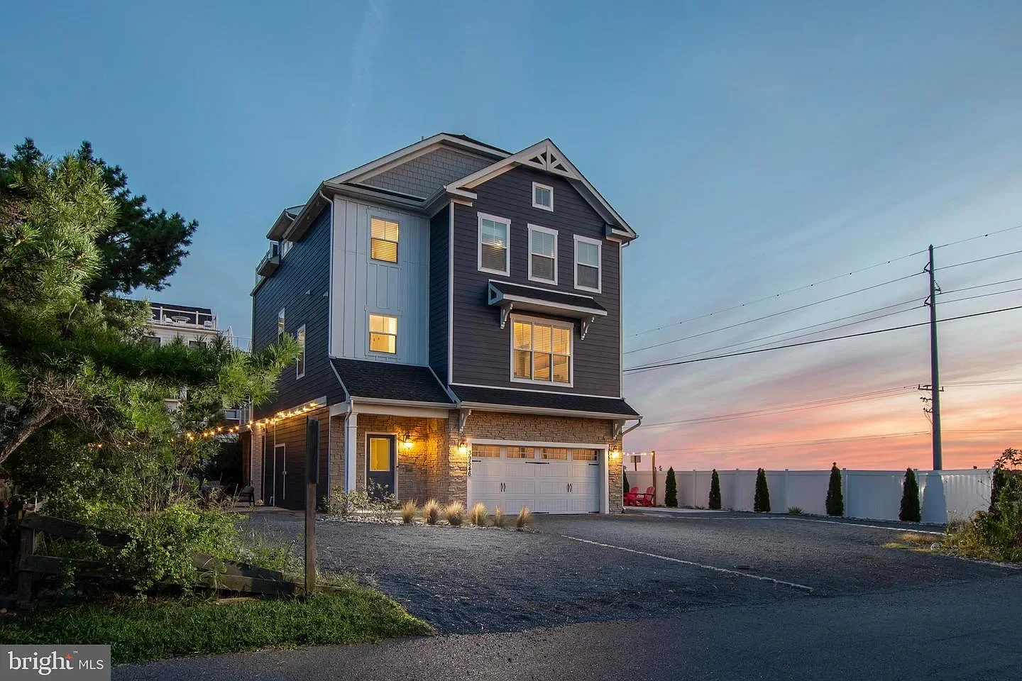 Three-story modern house with a garage, white fence, and landscaping at sunset.