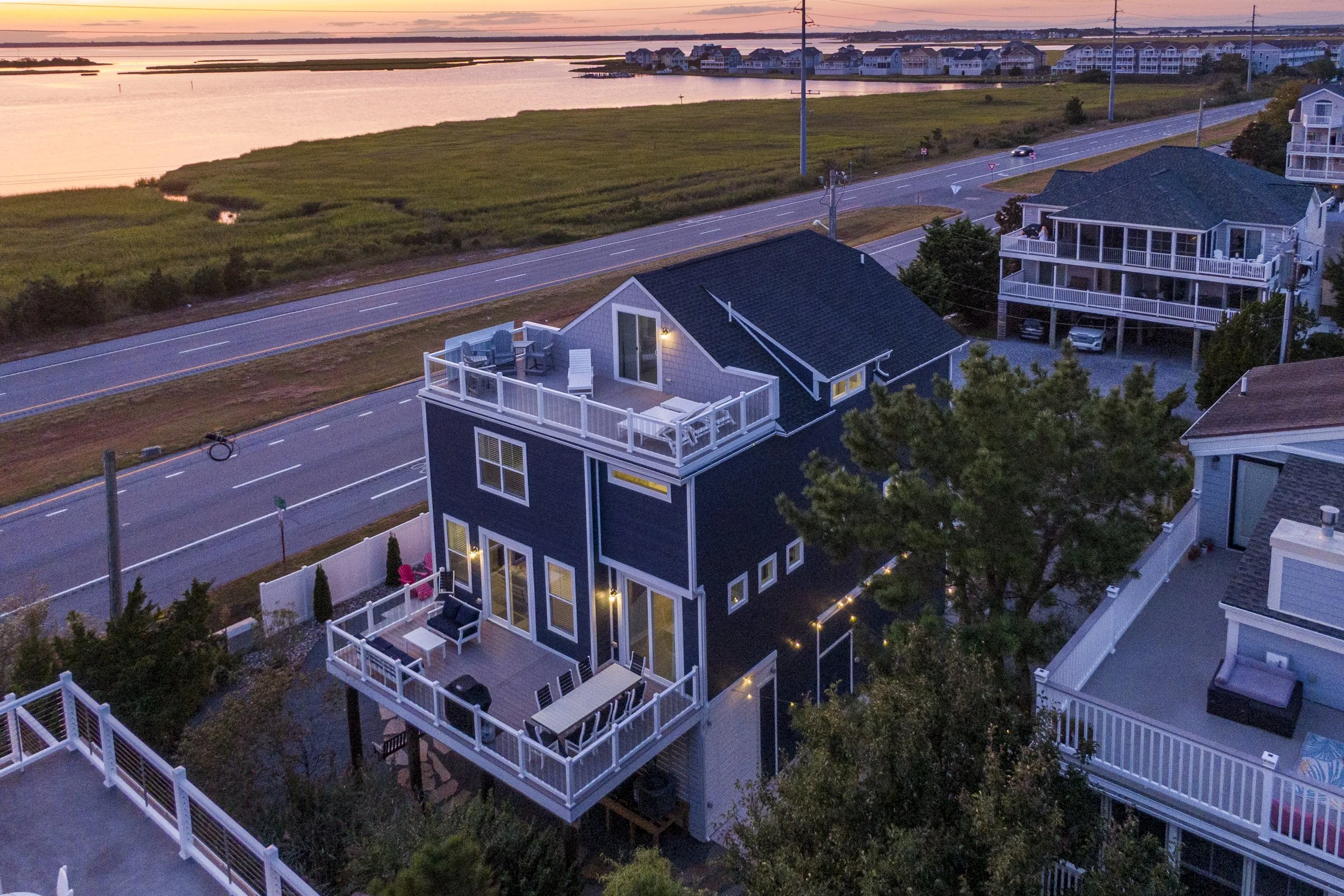 Aerial view of a blue multistory house with a large rooftop deck, white railings, outdoor furniture, and a lit outdoor space, near a road and a body of water during sunset.