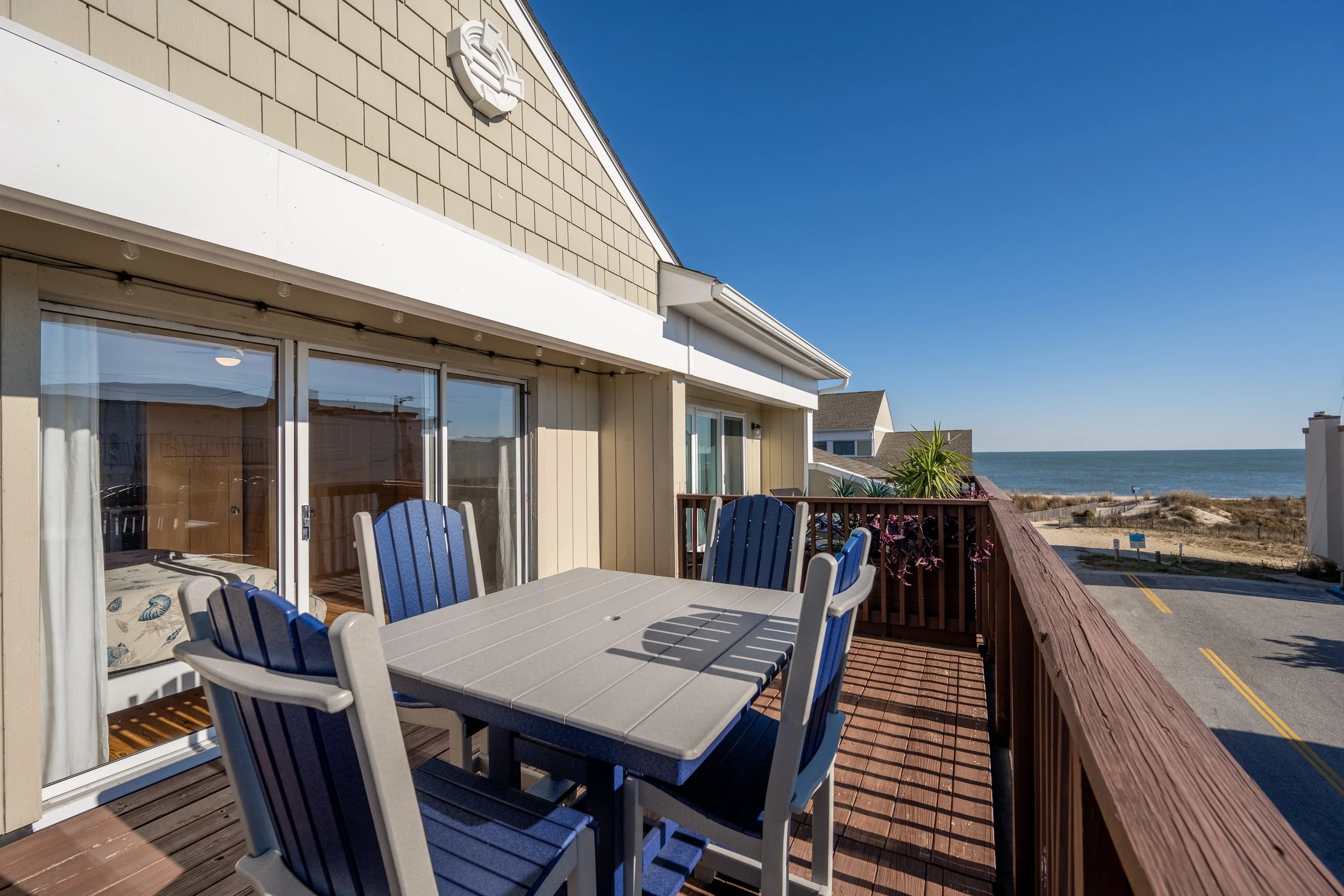 A balcony with outdoor furniture, including a table and chairs, overlooking the beach and ocean on a sunny day.