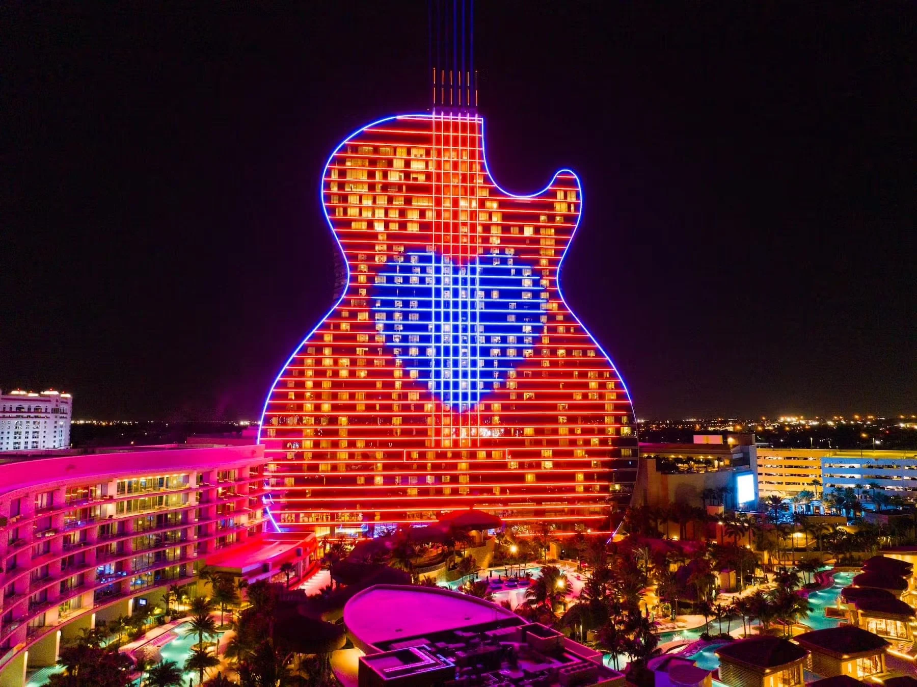 Nighttime view of a hotel shaped like a guitar with a neon heart outline, surrounded by a pool area and other buildings.