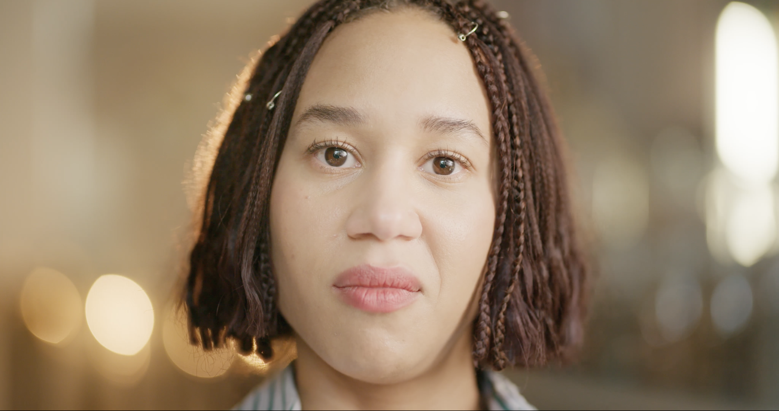 Close-up of a young woman with short, auburn braided hair and hazel eyes, looking into the camera, with a blurred background of lights.