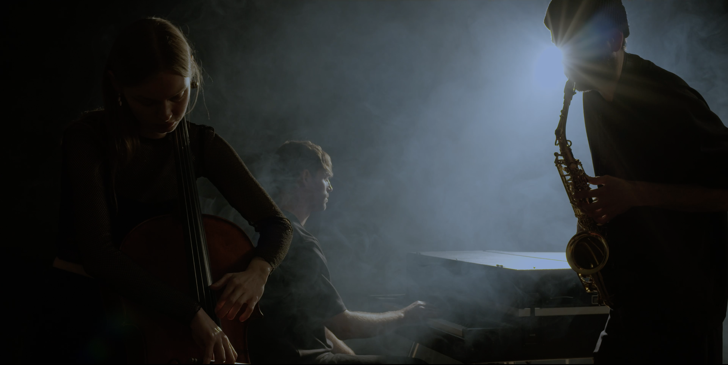 Three musicians are playing in a dimly lit, smoky room, with a female cellist, a male pianist, and a male saxophonist. A bright light illuminates them all from behind.