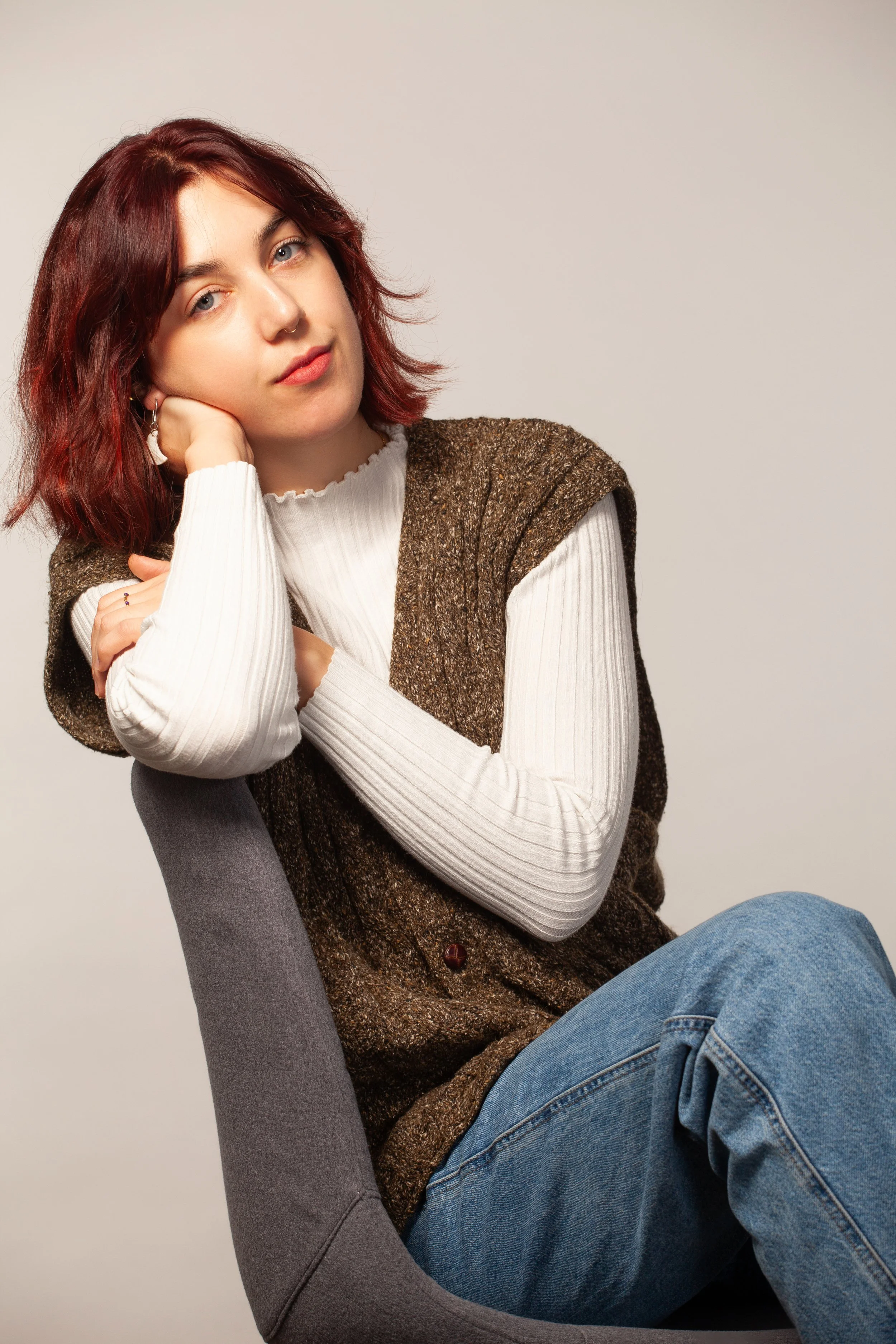 A woman with red hair, blue eyes, wearing a white ribbed sweater and brown vest, sitting on a gray modern chair, posing with her head resting on her hand against a plain light background.