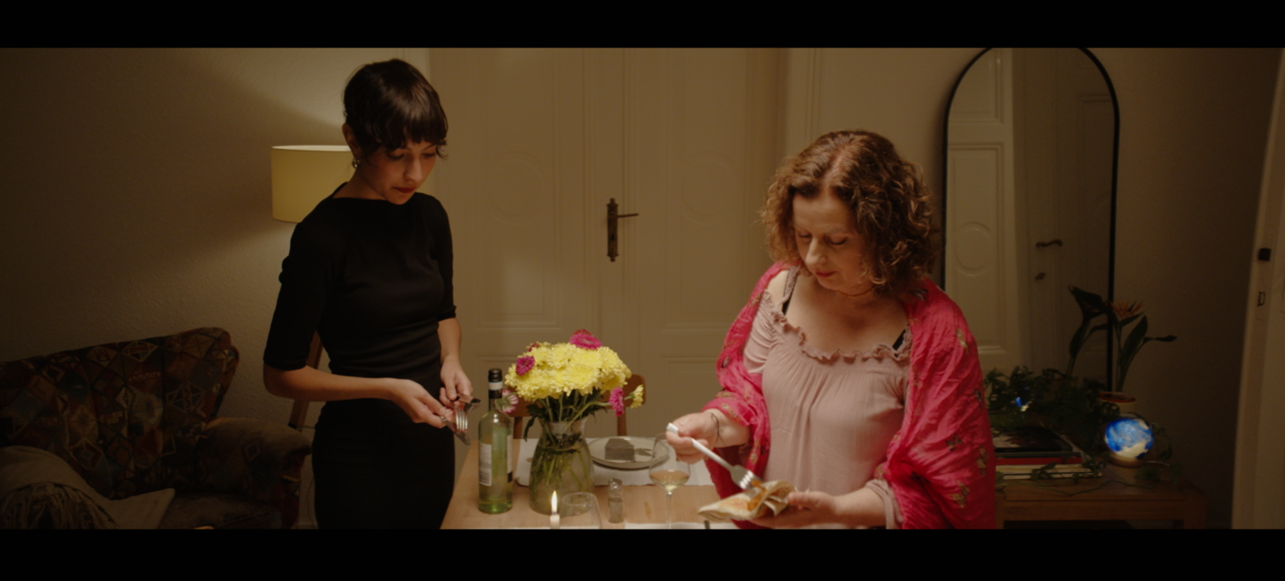 Two women clearing a dining table with a vase of yellow and pink flowers, a bottle of wine, and wine glasses in a warmly lit room.