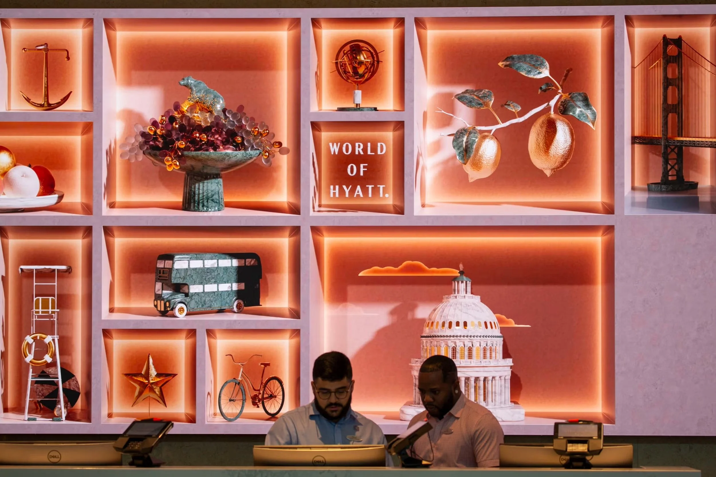 Decorative wall shelves with decorations at the Hyatt Hotel, including ornaments, a boat model, a globe, a fruit branch, a double-decker bus, a bicycle, a star, a Ferris wheel, and a Capitol Building replica, with two staff members at the reception desk.