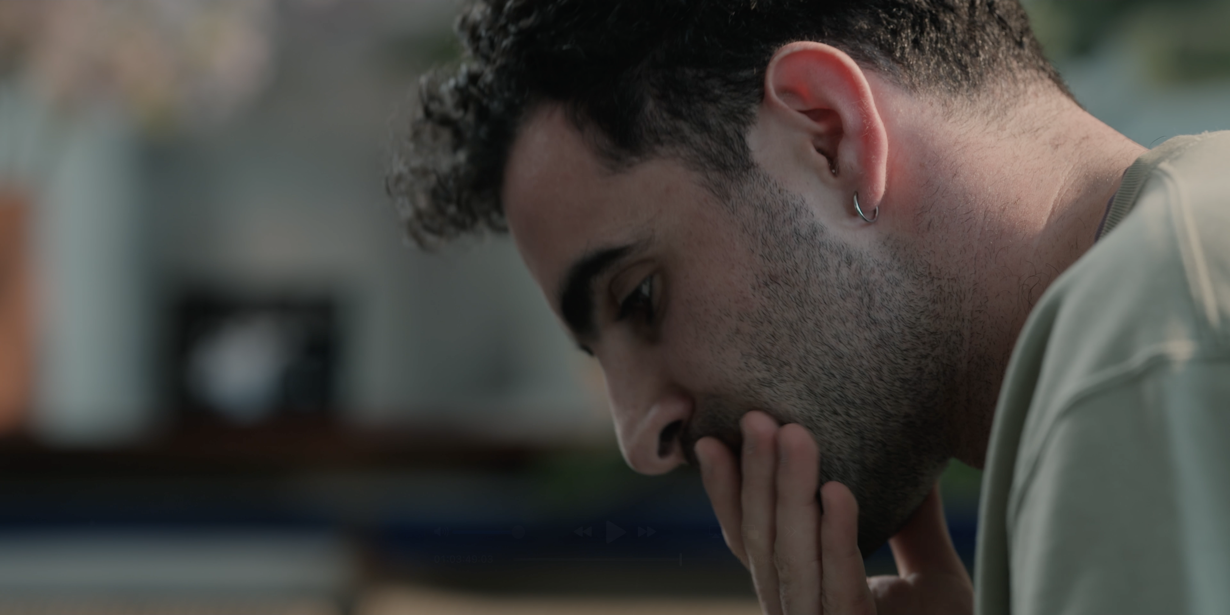 A young man with dark hair, a beard, and an earring is covering his mouth with his hand, looking down thoughtfully.