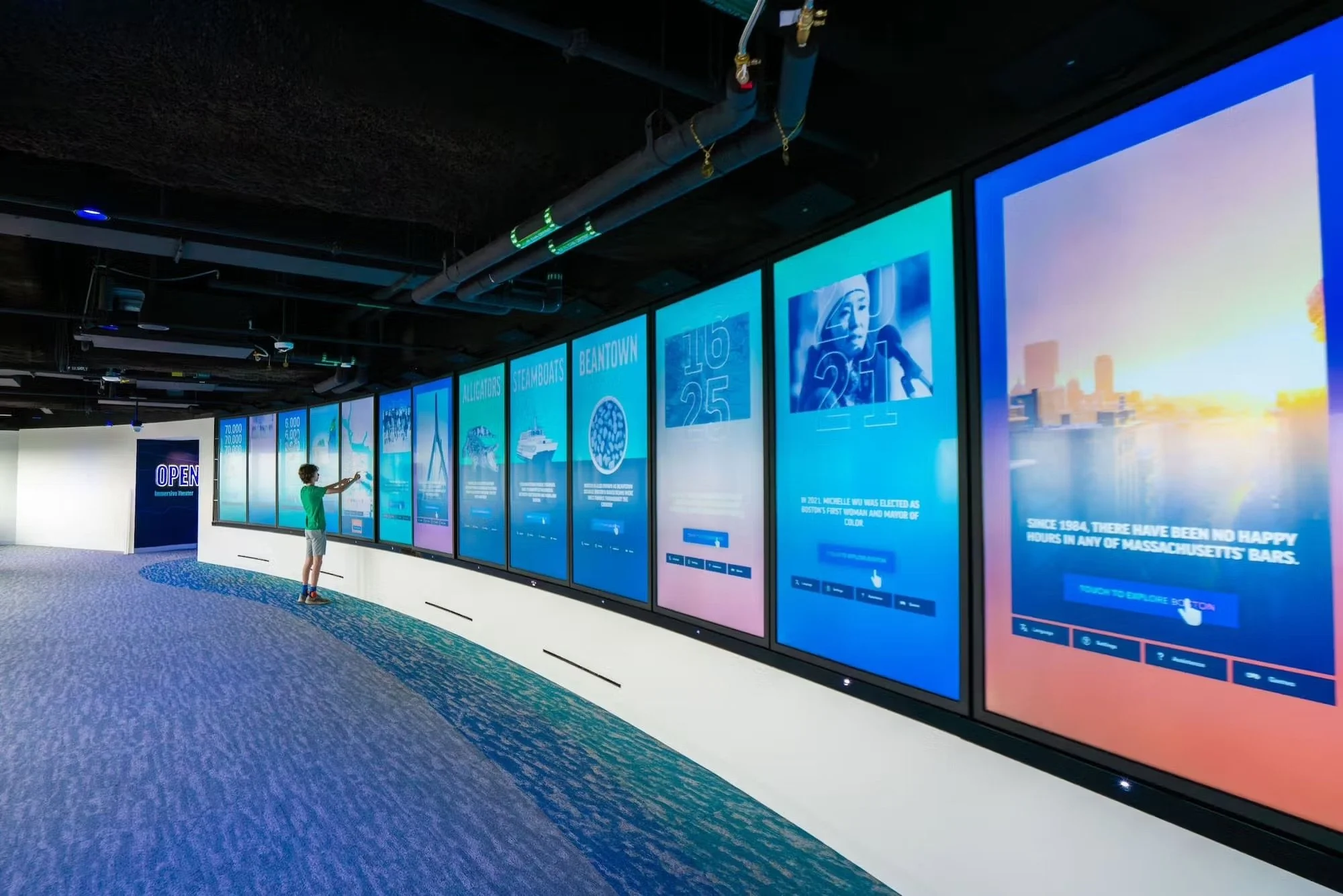 A young person in a green shirt and shorts interacts with large digital screens in an indoor exhibit space, with information and images displayed on the screens.