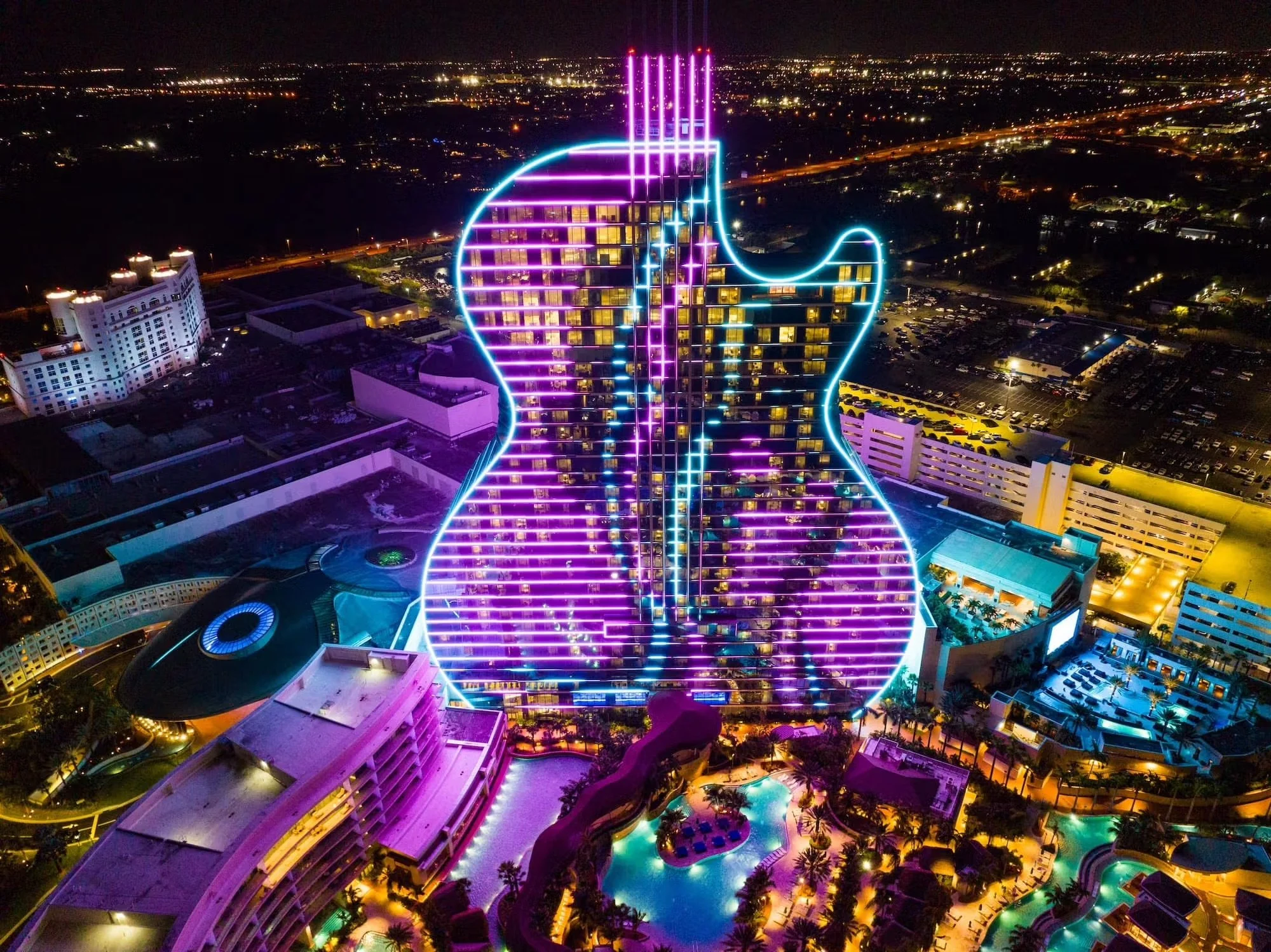 Aerial nighttime view of a hotel shaped like a guitar, outlined with bright neon purple and blue lights, with a surrounding pool area and cityscape in the background.
