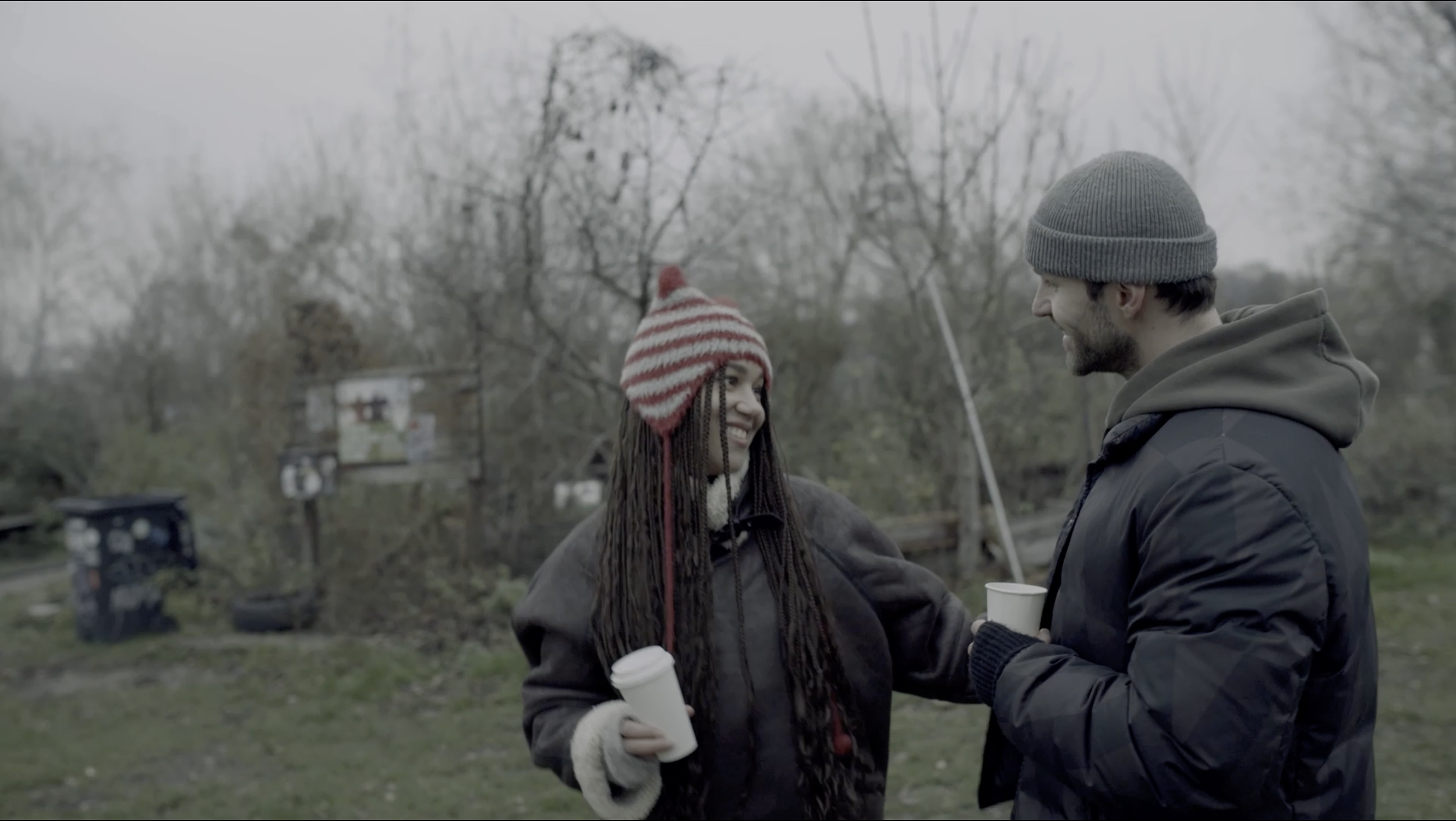 A man and woman talking outdoors in a park on a cloudy day, both holding white cups, dressed warmly in jackets and beanies, smiling at each other.