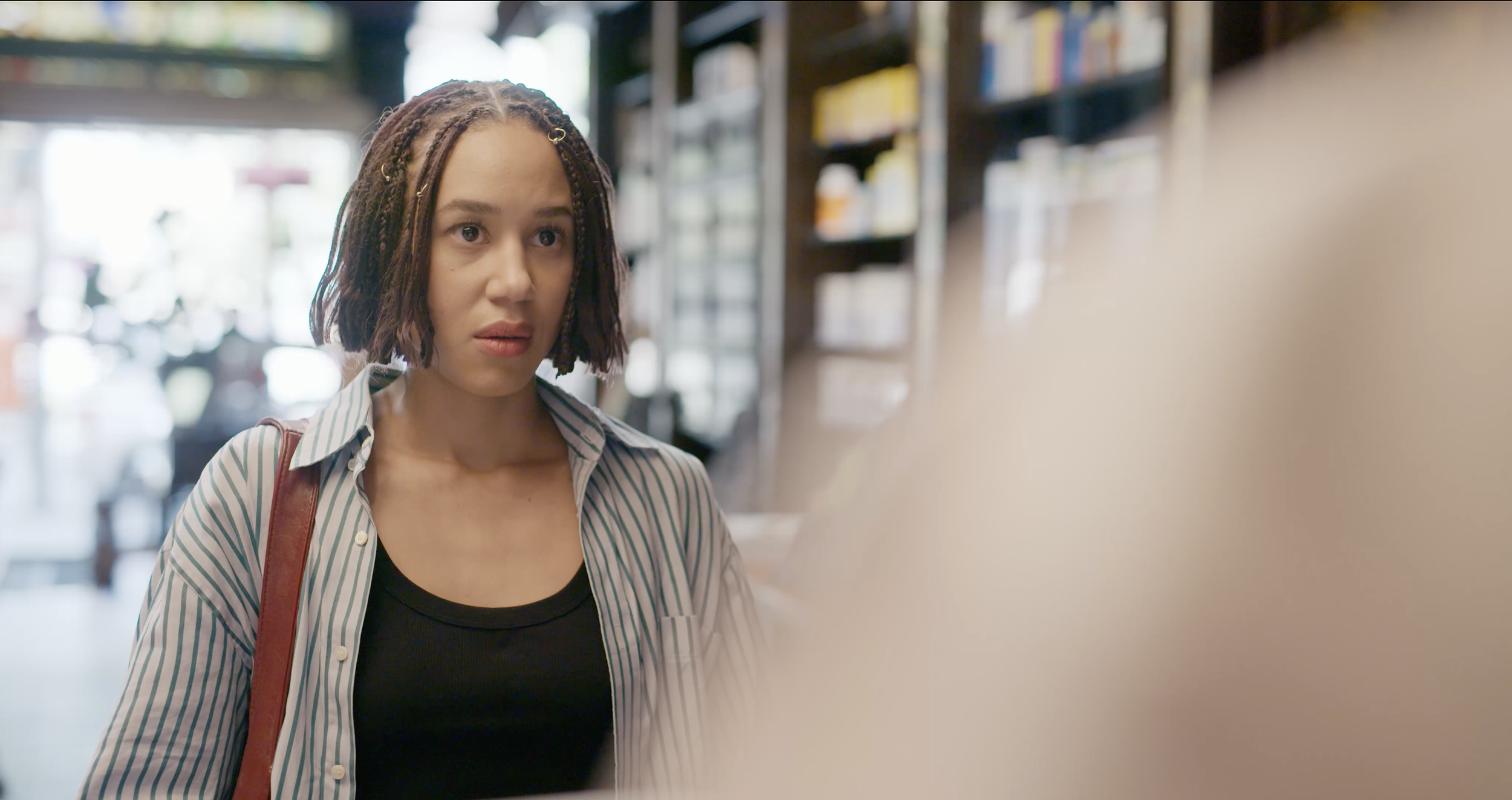 Woman with short braided hair, wearing a black shirt and striped blazer, looking intently at a pharmacist in a pharmacy.