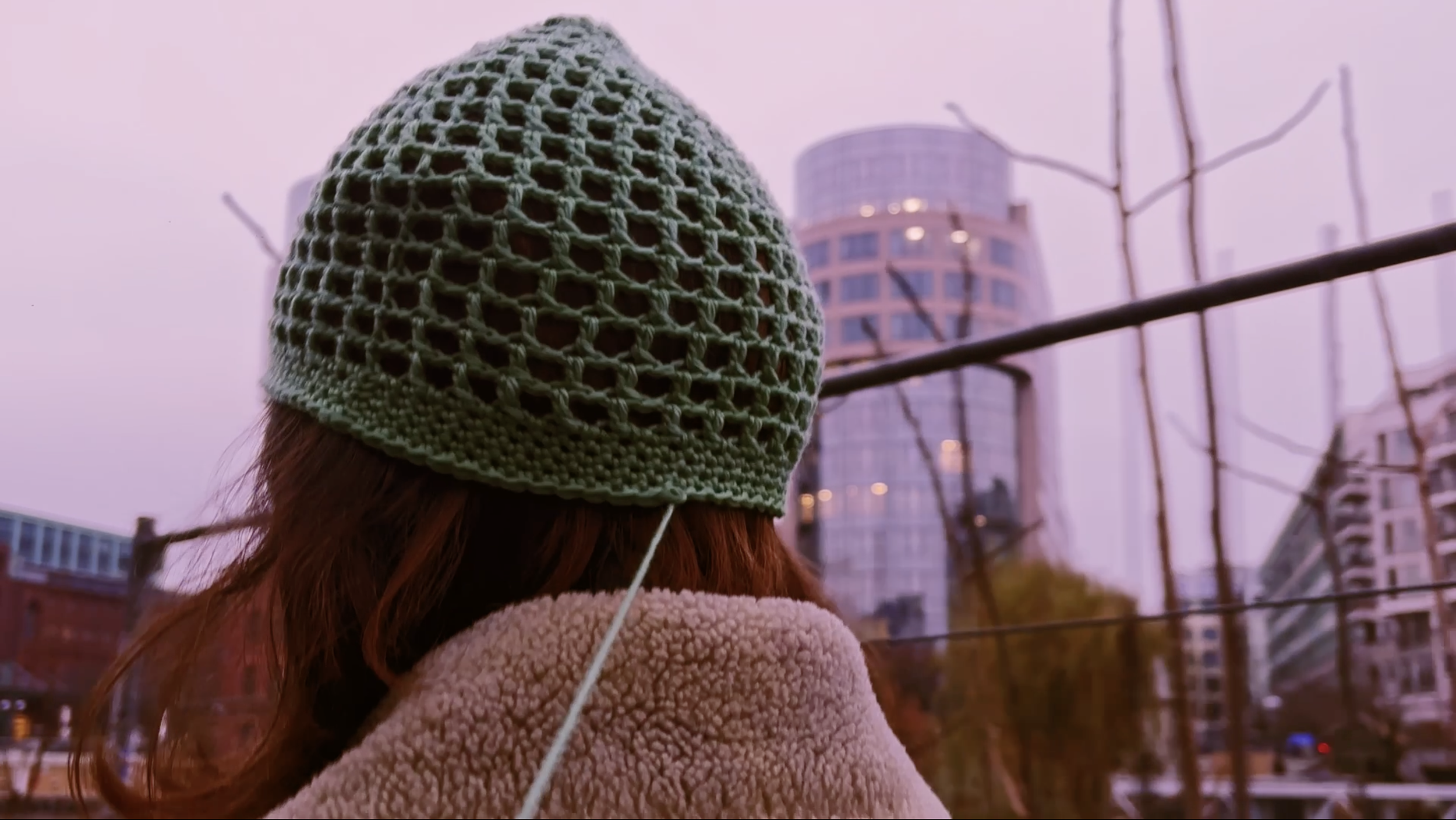 Person wearing a green crochet beanie and a beige fleece coat looking at city buildings and trees at sunset.