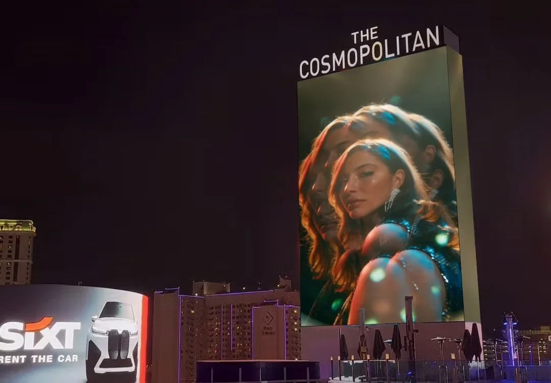 Large digital billboard on a building display magazine cover with a woman with long hair, wearing an elegant dress and earrings, with moody lighting and reflection effects on her face.