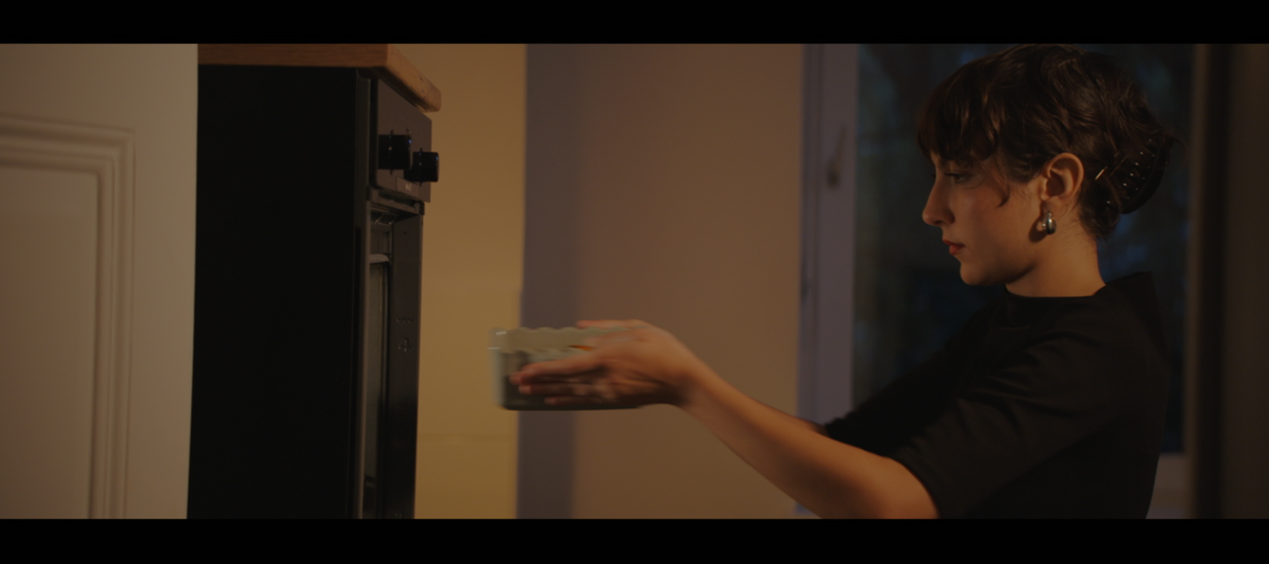 A woman with short dark hair, wearing earrings and a black top, is placing a dish into an oven.