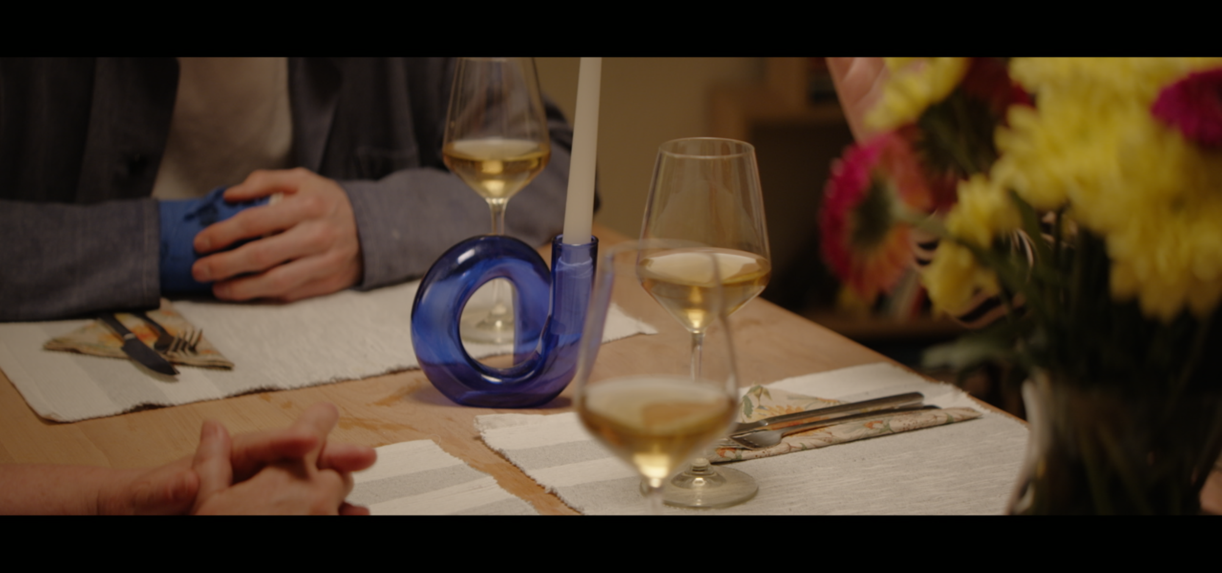 A dining table set with three glasses of white wine, a blue glass candle holder with a white candle, and a vase with yellow and pink flowers. A person with a blue cast on their wrist is seen with hands clasped, sitting at the table.