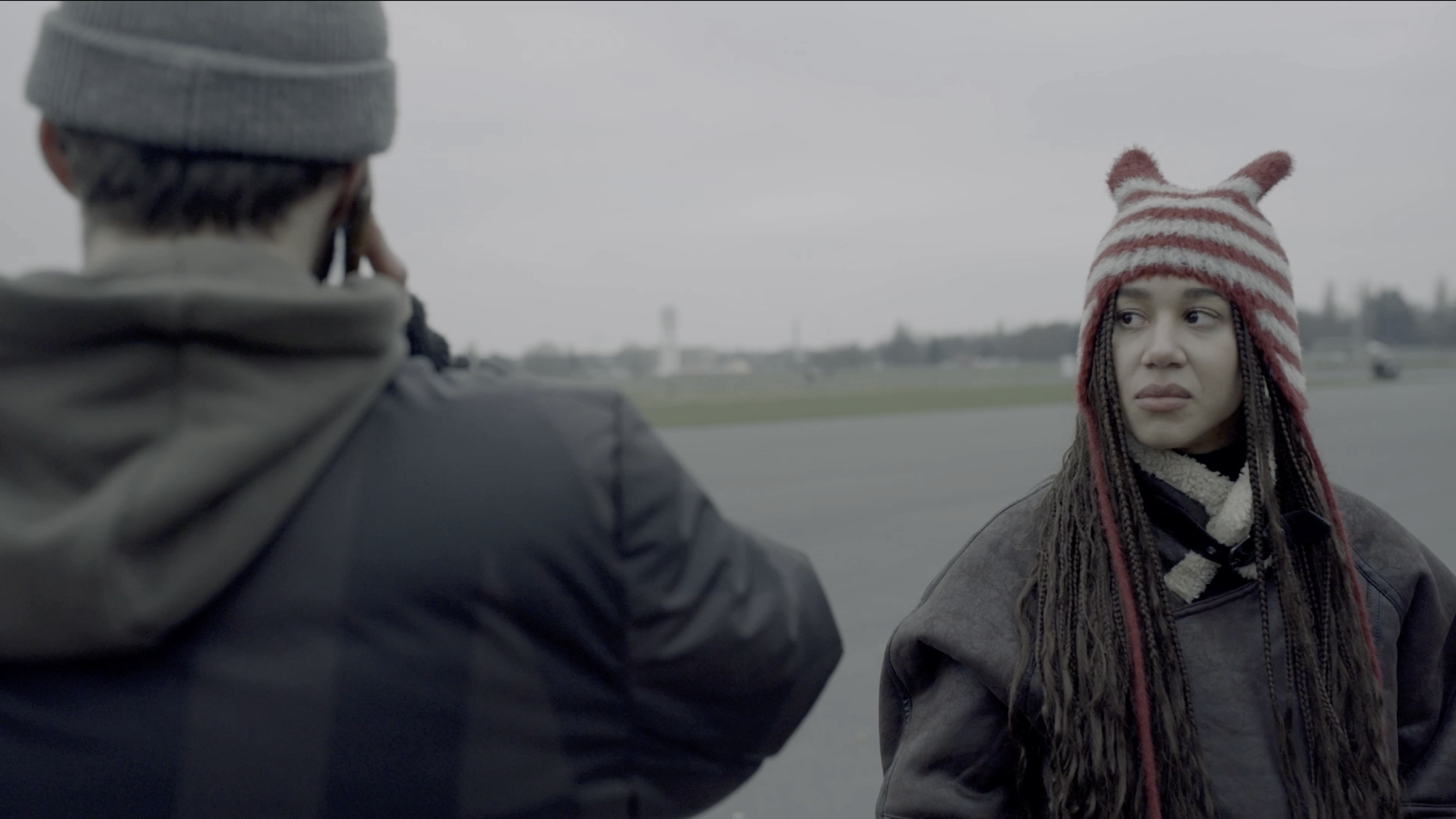 On an outdoor, overcast day, a man with a grey beanie taking a phone call while a woman with a red and white striped knit hat watches him. The woman has long braids and is wearing a dark jacket.