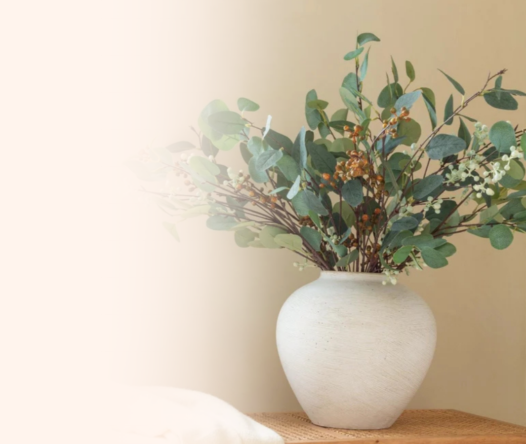 A white ceramic vase filled with green leafy branches and small white and brown flowers on a wooden surface against a beige wall.