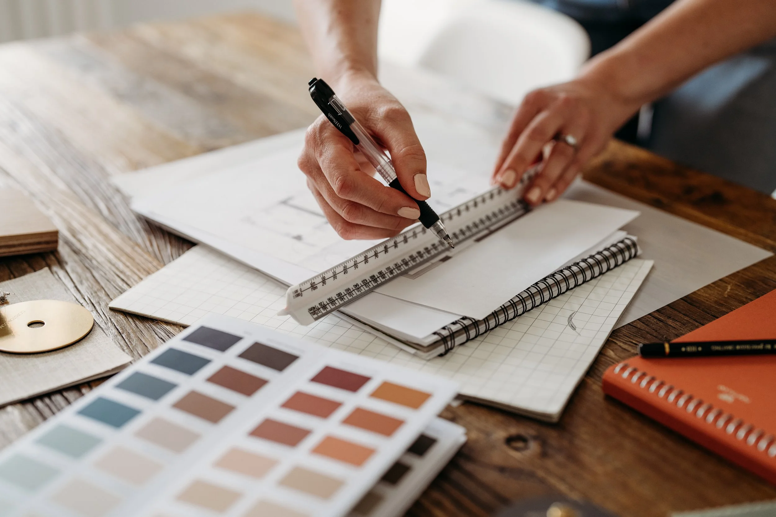 An interior desiggner is drawing a design on paper with a black pen and a scale ruler on a wooden table surrounded by various notebooks, color swatches, and paper materials.