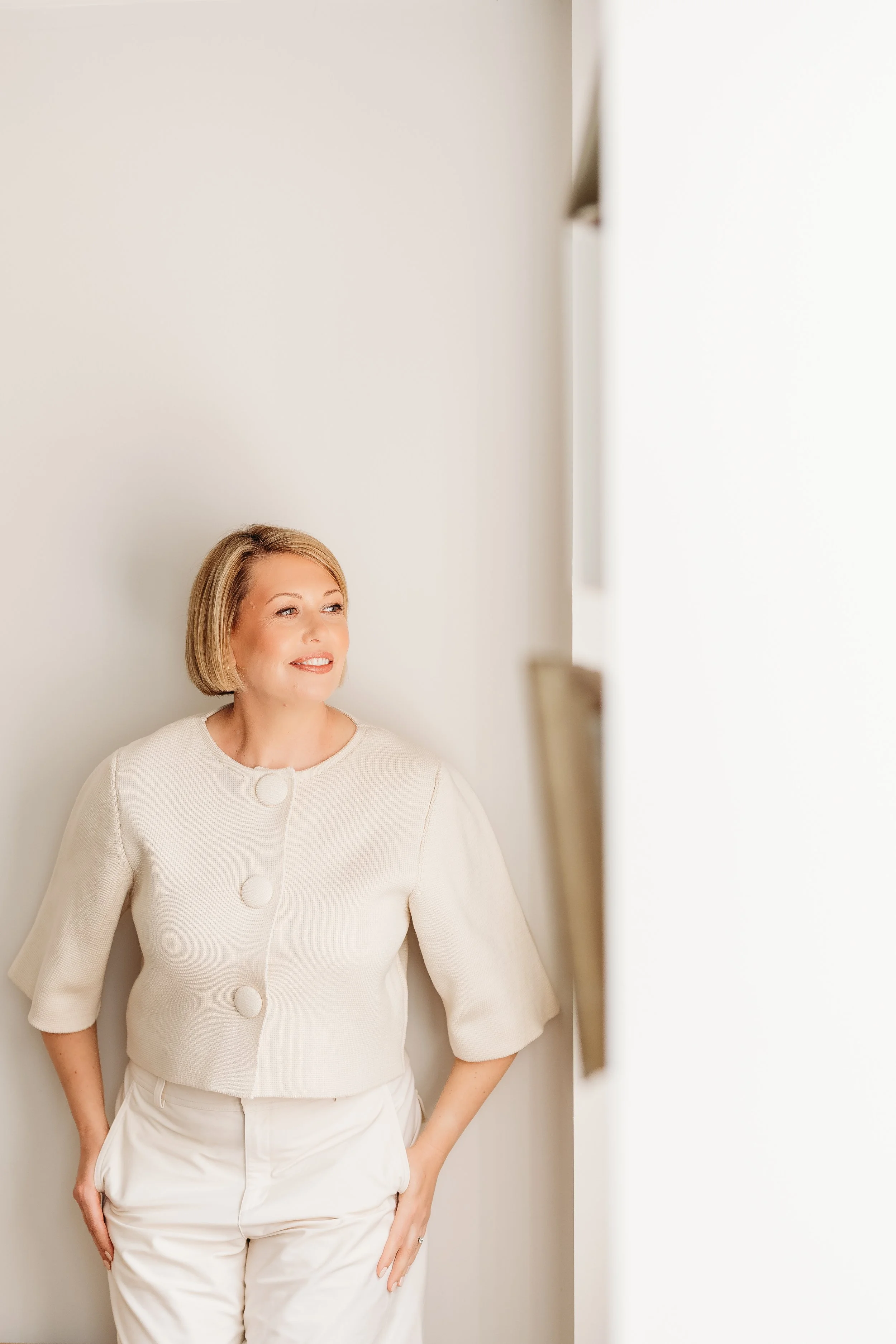 An interior designer smiling and looking to her left, wearing a cream-colored jacket and pants, standing in front of a plain white wall.