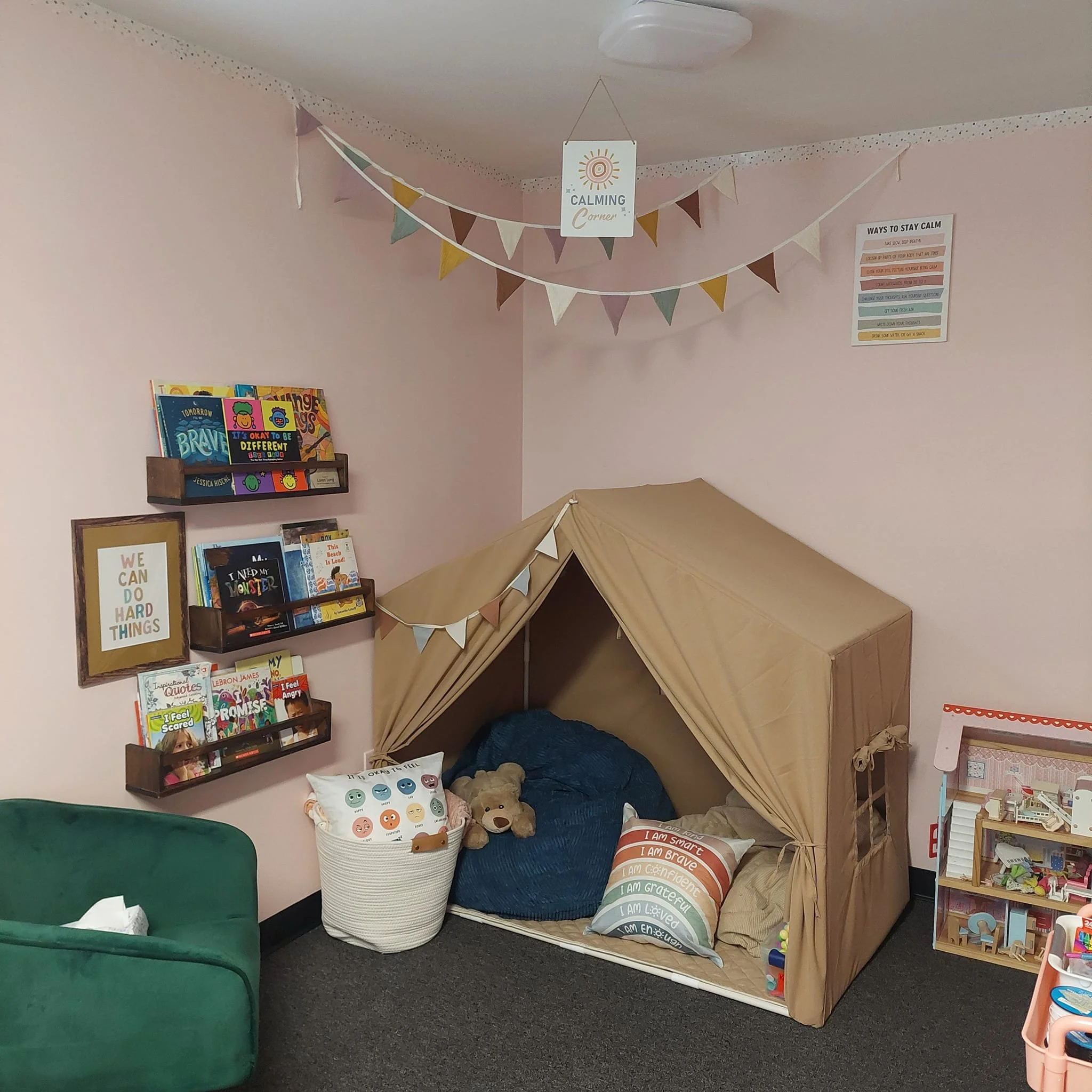 Children's reading nook with pink walls, a small tent with bedding and plush toy, bookshelf with children's books, decorative bunting and a calming sign hanging from the ceiling, colorful poster on the wall, and a small green armchair in the corner.