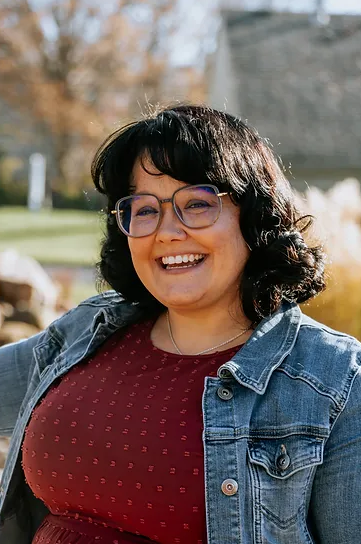 A woman with black hair, glasses, and a wide smile, wearing a maroon shirt and a denim jacket, standing outdoors on a sunny day.