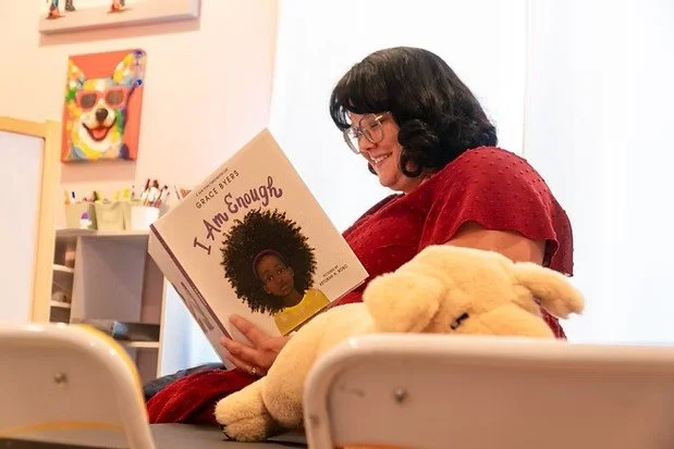 Woman with glasses reading a children's picture book titled "I Am Enough" by Grace Byers, sitting with a stuffed animal on her lap, in a cozy room with colorful wall art behind her.