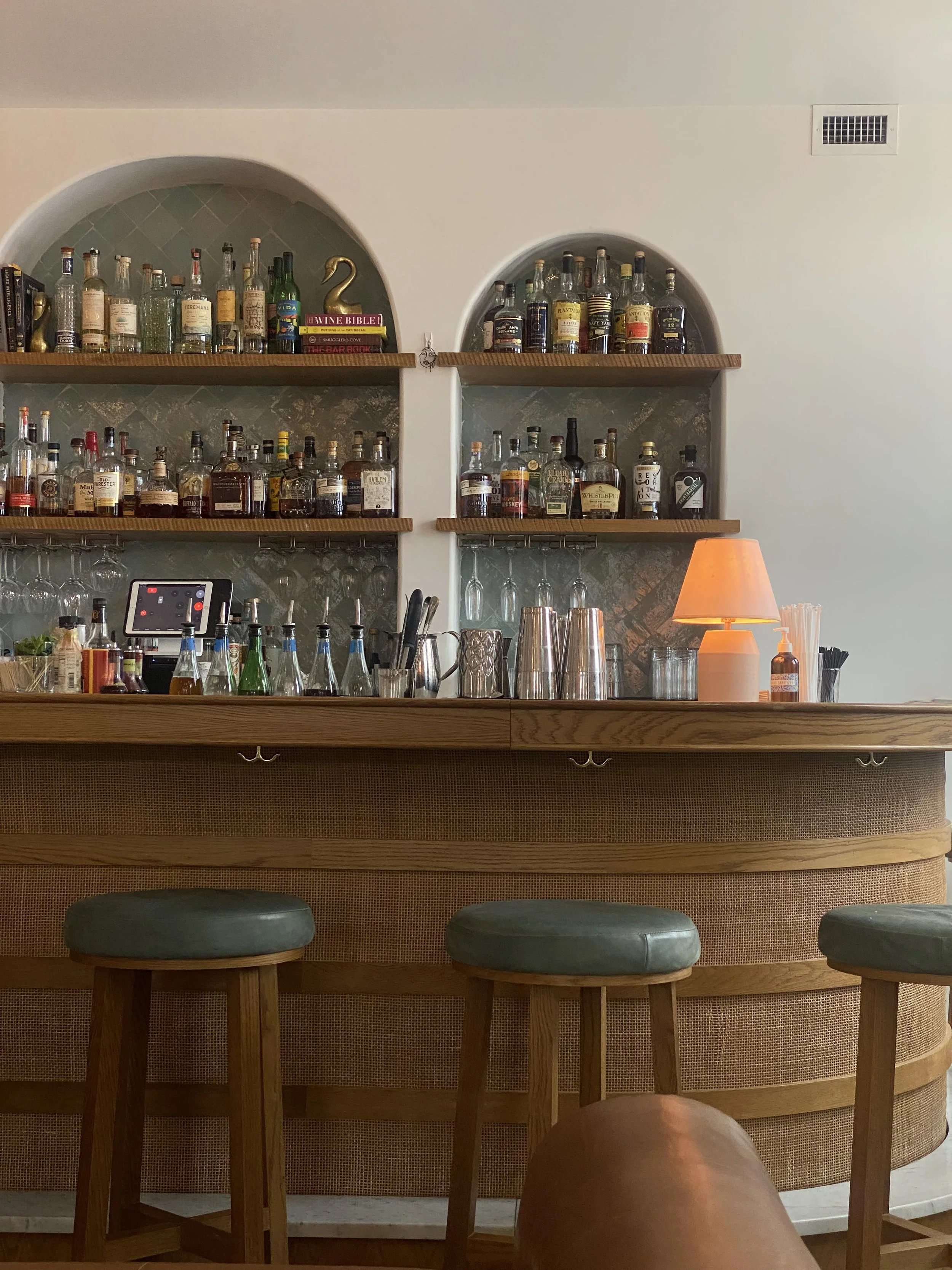 View of a stylish bar with shelves of liquor bottles, bar tools, and a small lamp, with three green bar stools in front.