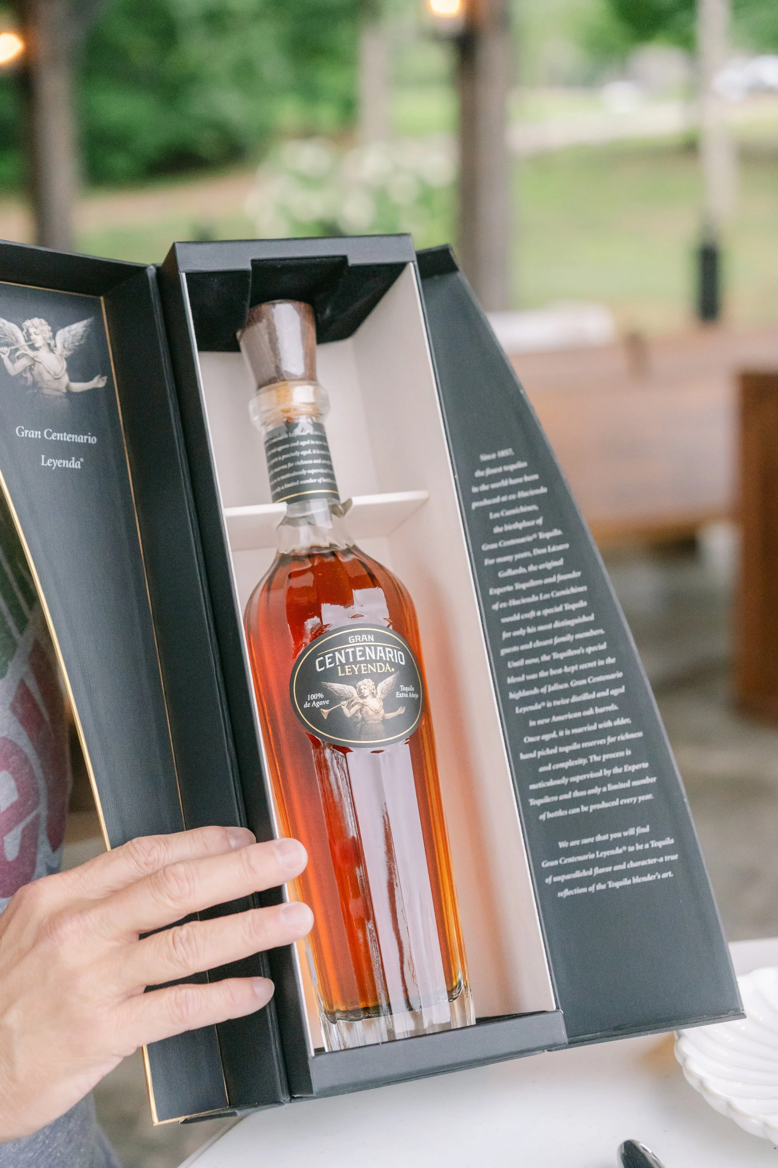 A person holding a boxed bottle of Gran Centenario Leyenda tequila, which is visible through the open box with informational text on the right side.