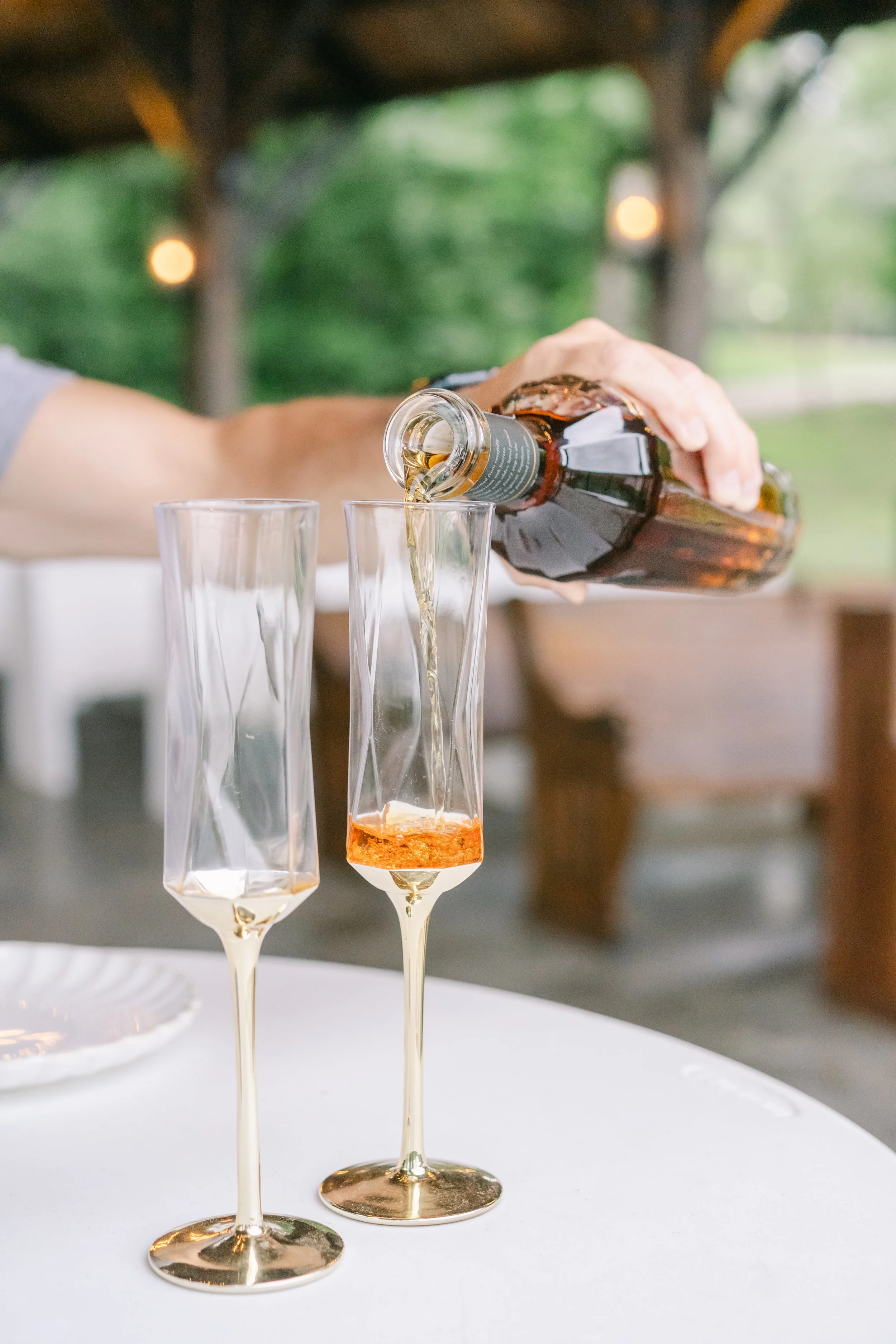Person pouring a drink into two champagne flutes at an outdoor gathering with a blurred green background.