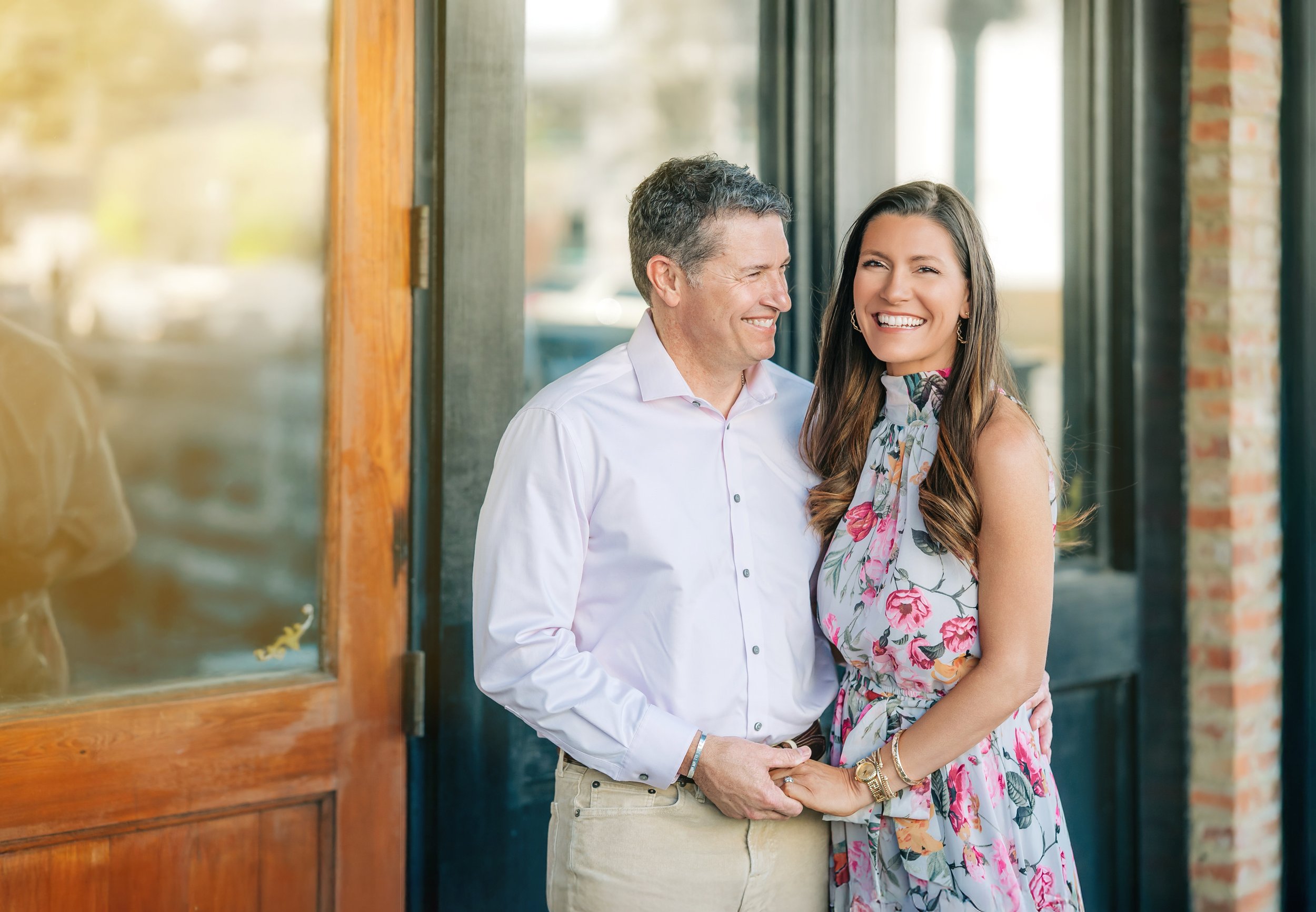 A middle-aged man and a young woman are smiling and holding hands, standing close to each other near a doorway with large glass windows, with a blurred background of a street and trees.