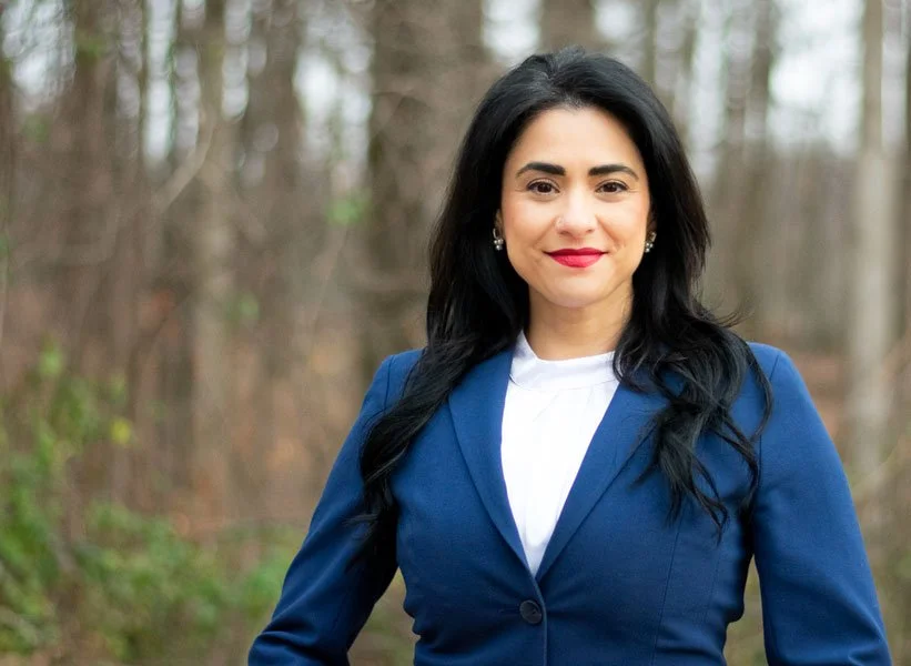 A woman with black hair, wearing a blue blazer and white blouse, standing outdoors in a wooded area with trees in the background.