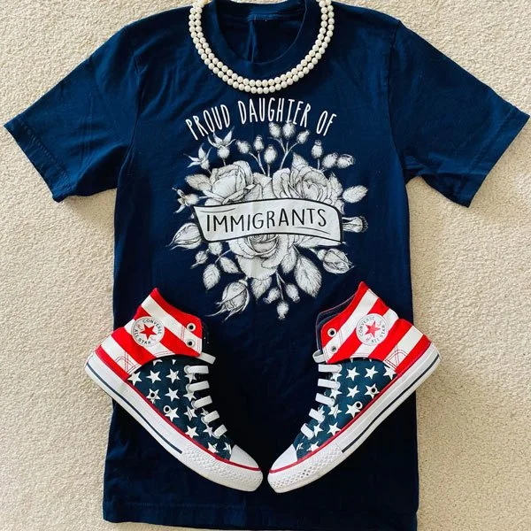 A navy blue T-shirt with a floral graphic and the text 'Proud Daughter of Immigrants', a pair of red, white, and blue high-top Converse sneakers with stars and stripes pattern, and a pearl necklace.