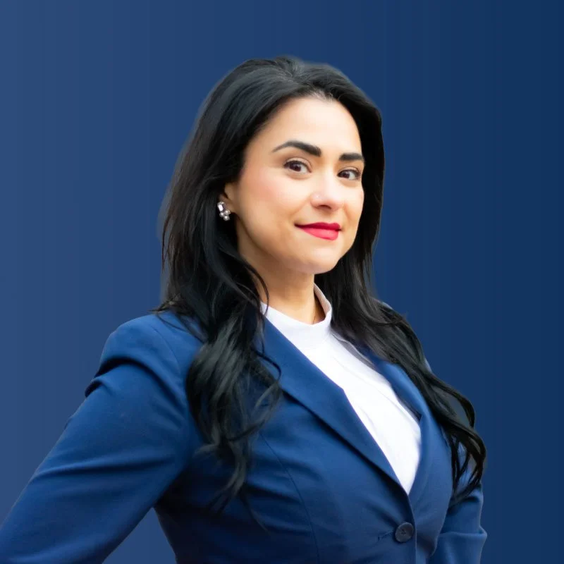 Professional woman with long black hair, wearing a blue blazer and pearl earrings, standing against a blue background.