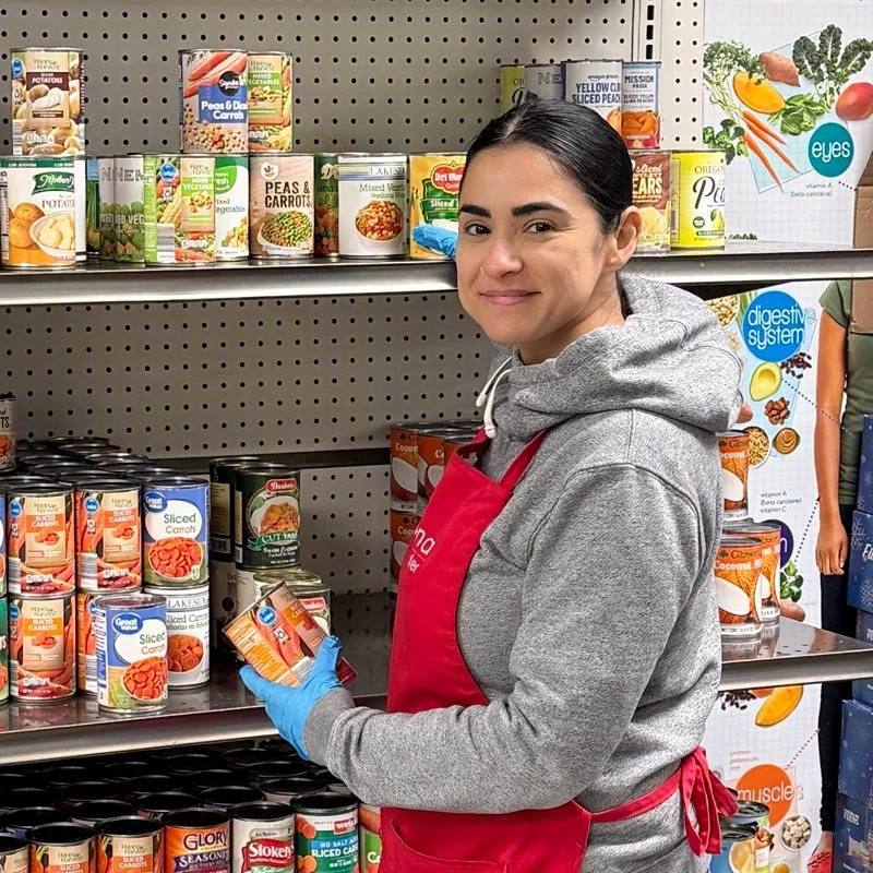 Woman wearing a gray hoodie, red apron, and blue gloves shopping for canned vegetables at a store aisle with canned carrots and peas.