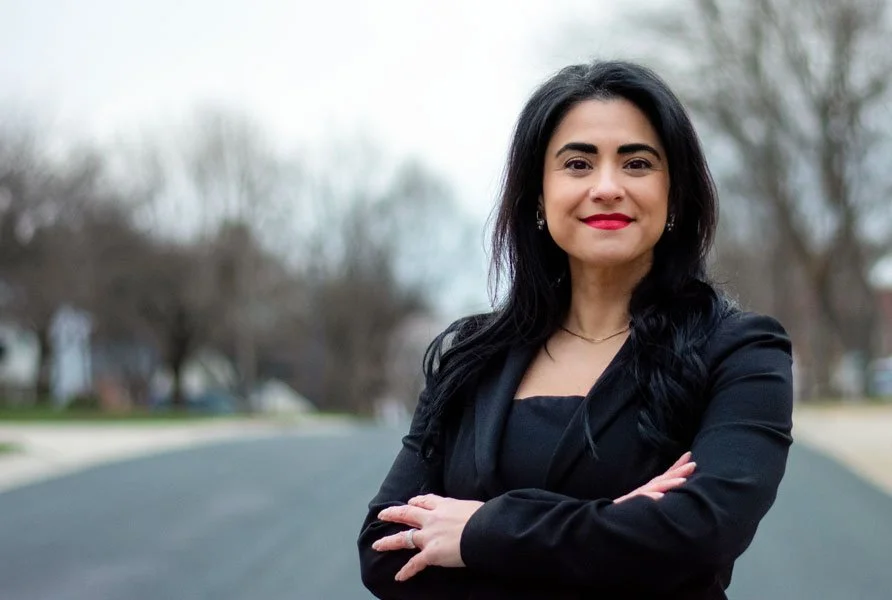A woman with black hair, wearing a black blazer and black blouse, standing outdoors in a residential area with trees in the background.